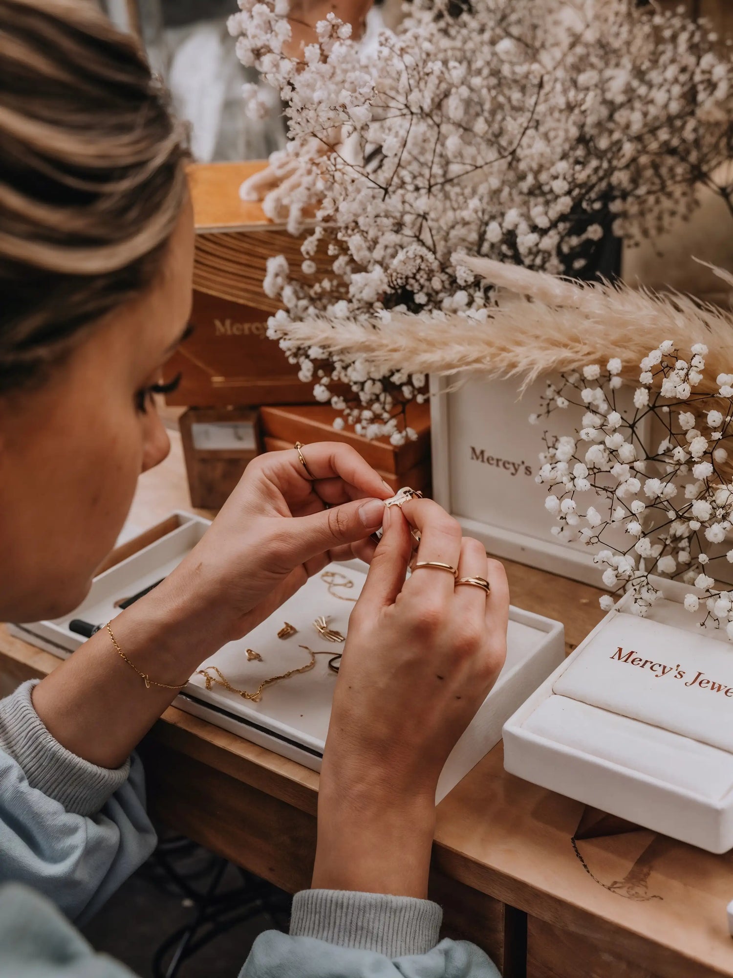 Hände fertigen Mercy’s Jewelry Goldschmuck handgemacht in einer Werkstatt, mit Verpackungen und Blumendeko im Hintergrund