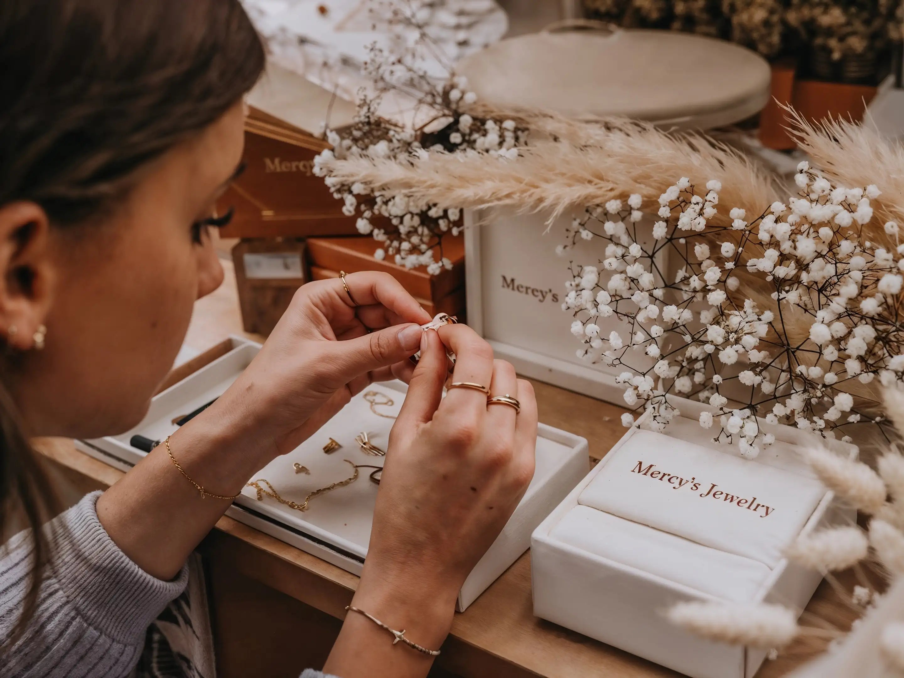 Nahaufnahme einer Frau, die handgefertigten Schmuck von Mercy’s Jewelry bearbeitet – goldene Ringe und Armbänder auf Schmucktablett