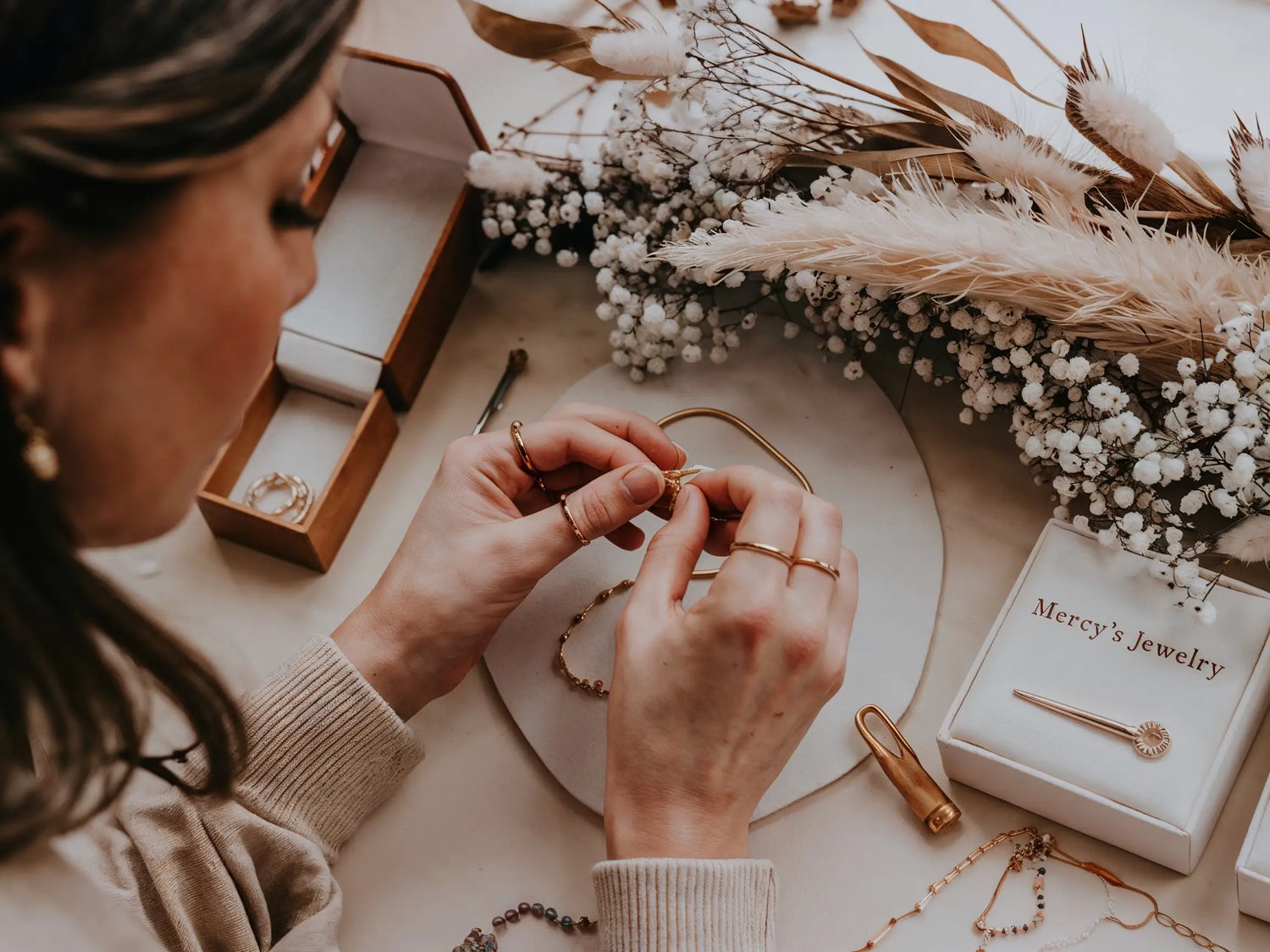 Hände arbeiten an Mercy’s Jewelry Schmuckstück in Handarbeit, mit Werkzeugen und Blumenarrangement im Hintergrund