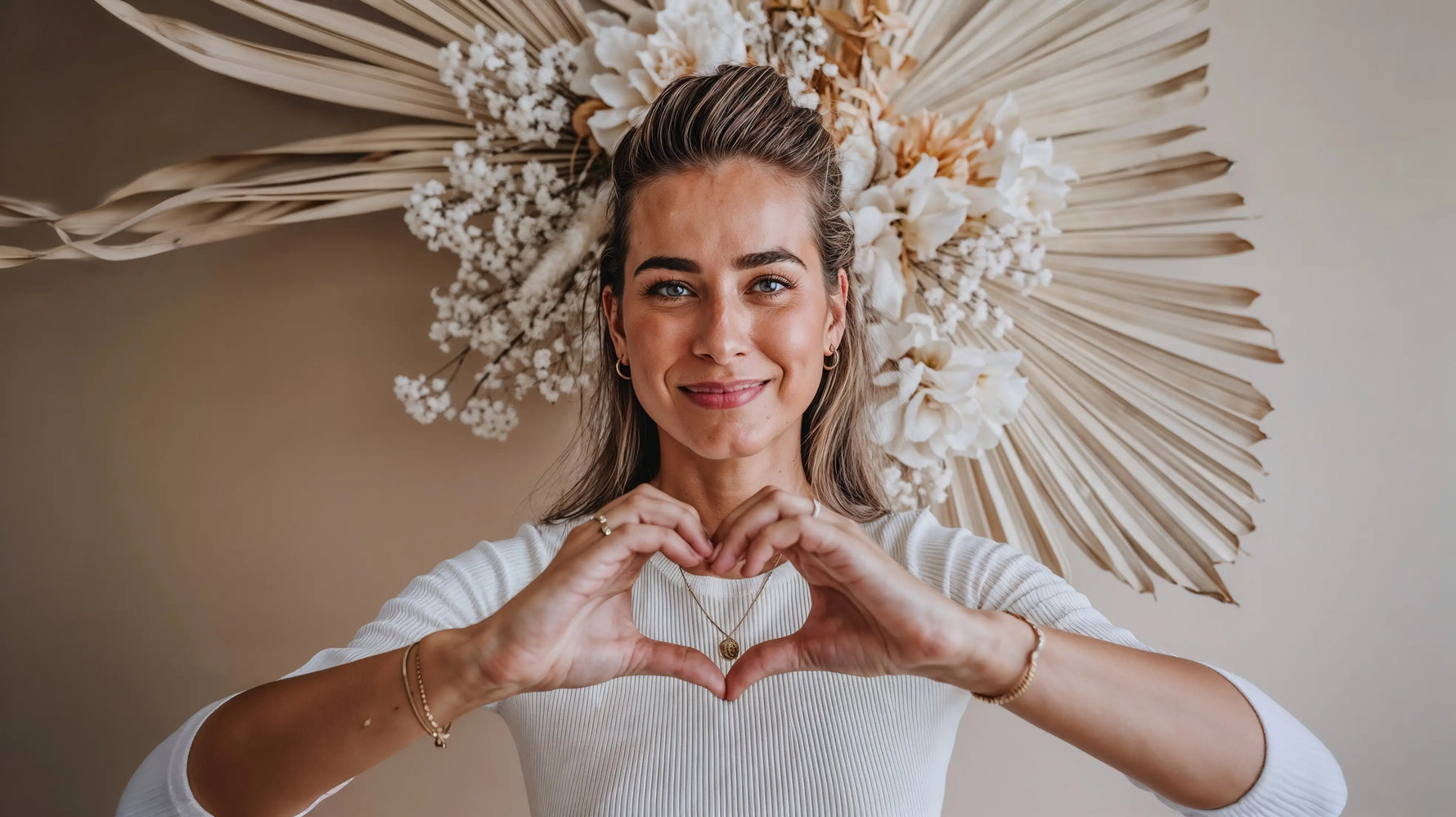 Model mit Mercy’s Jewelry Schmuck – goldene Halsketten, Armband und Ohrringe – formt mit den Händen ein Herz vor dekorativen Trockenblumen
