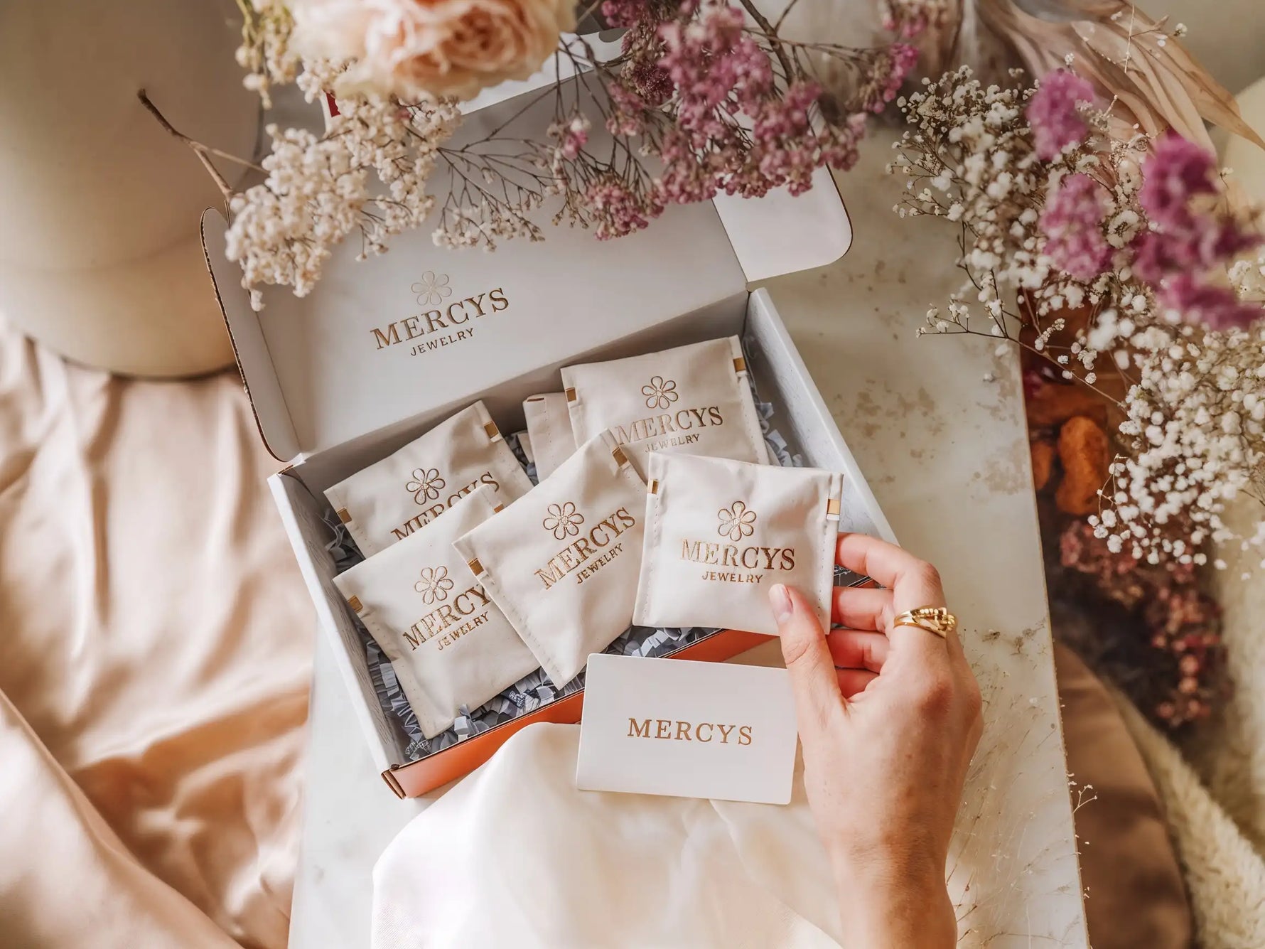 Offene Box mit mehreren Mercy’s Jewelry Schmuckpouches, dekoriert mit Blumen, eine Hand hält einen Pouch