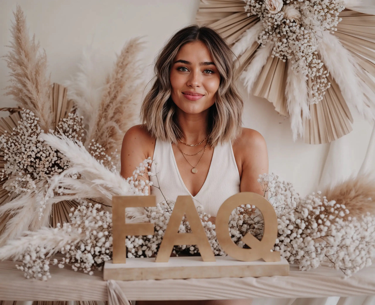 Model mit kurzem Haar trägt goldene Halsketten, sitzt vor Trockenblumenarrangement, davor große goldene Buchstaben FAQ