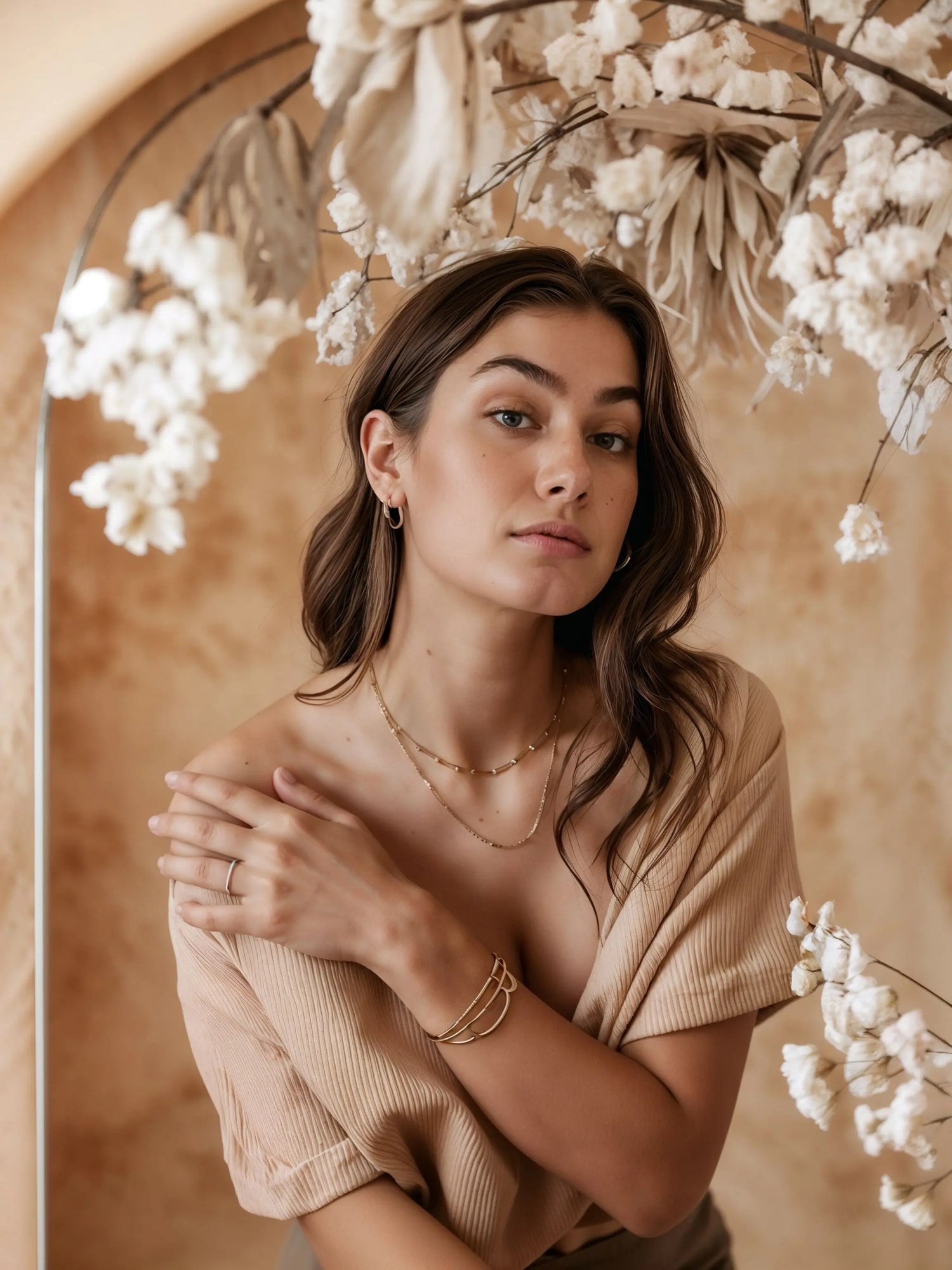 Model steht vor Blumendeko und trägt Mercy’s Jewelry Schmuck – goldene Halsketten im Layering-Look, Armband und kleine Creolen