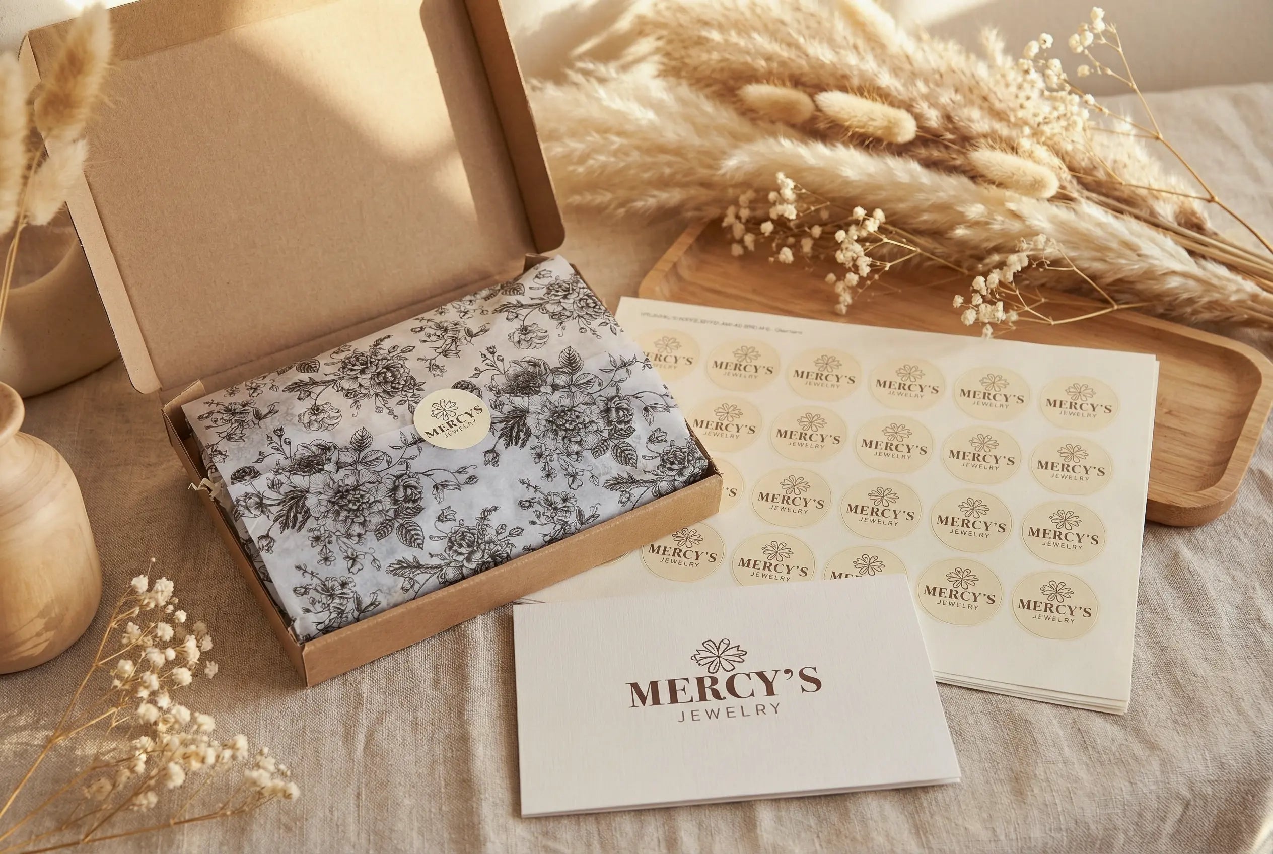 Offene Mercy’s Jewelry Versandbox mit floralem Seidenpapier und Stickerbogen auf einem Holztisch, dekoriert mit Pampasgras und Baby’s Breath im warmen Licht.