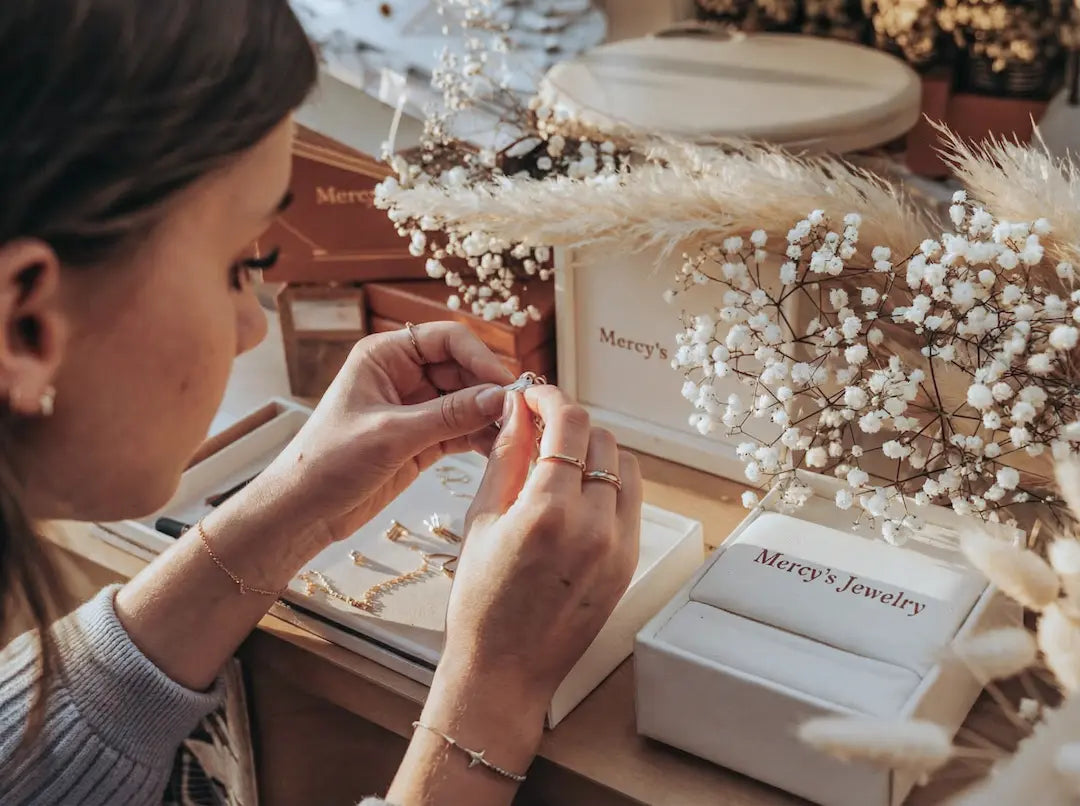 Eine Frau betrachtet filigranen Goldschmuck von MERCYS Jewelry in einem ästhetischen Setting mit Trockenblumen und edler Markenverpackung auf bymercy.de.