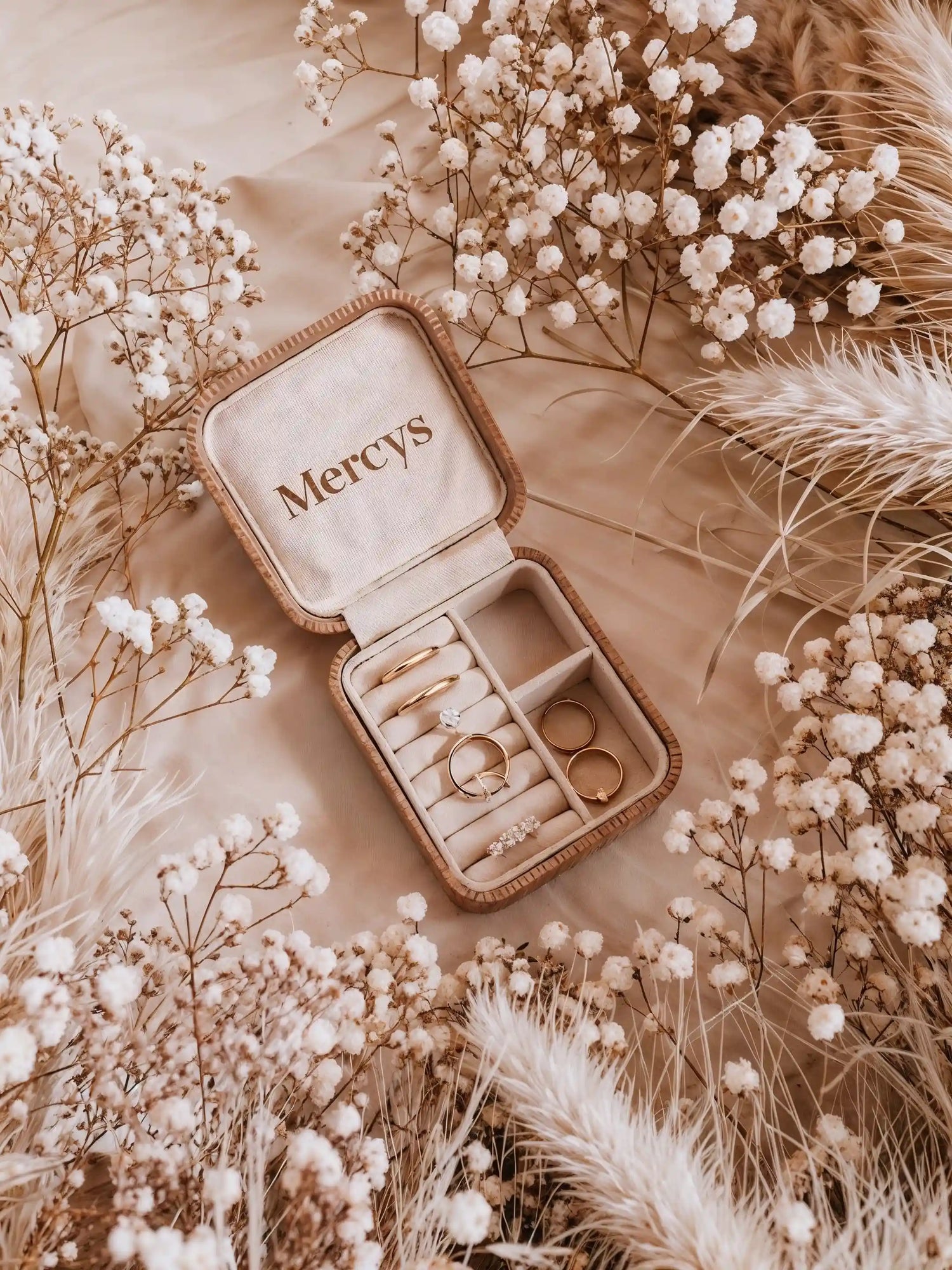 Schmuckkästchen von Mercy’s Jewelry mit goldenen Ringen auf beigem Stoff, dekoriert mit Trockenblumen – Schmuckpflegeguide zur sicheren Aufbewahrung von Schmuck