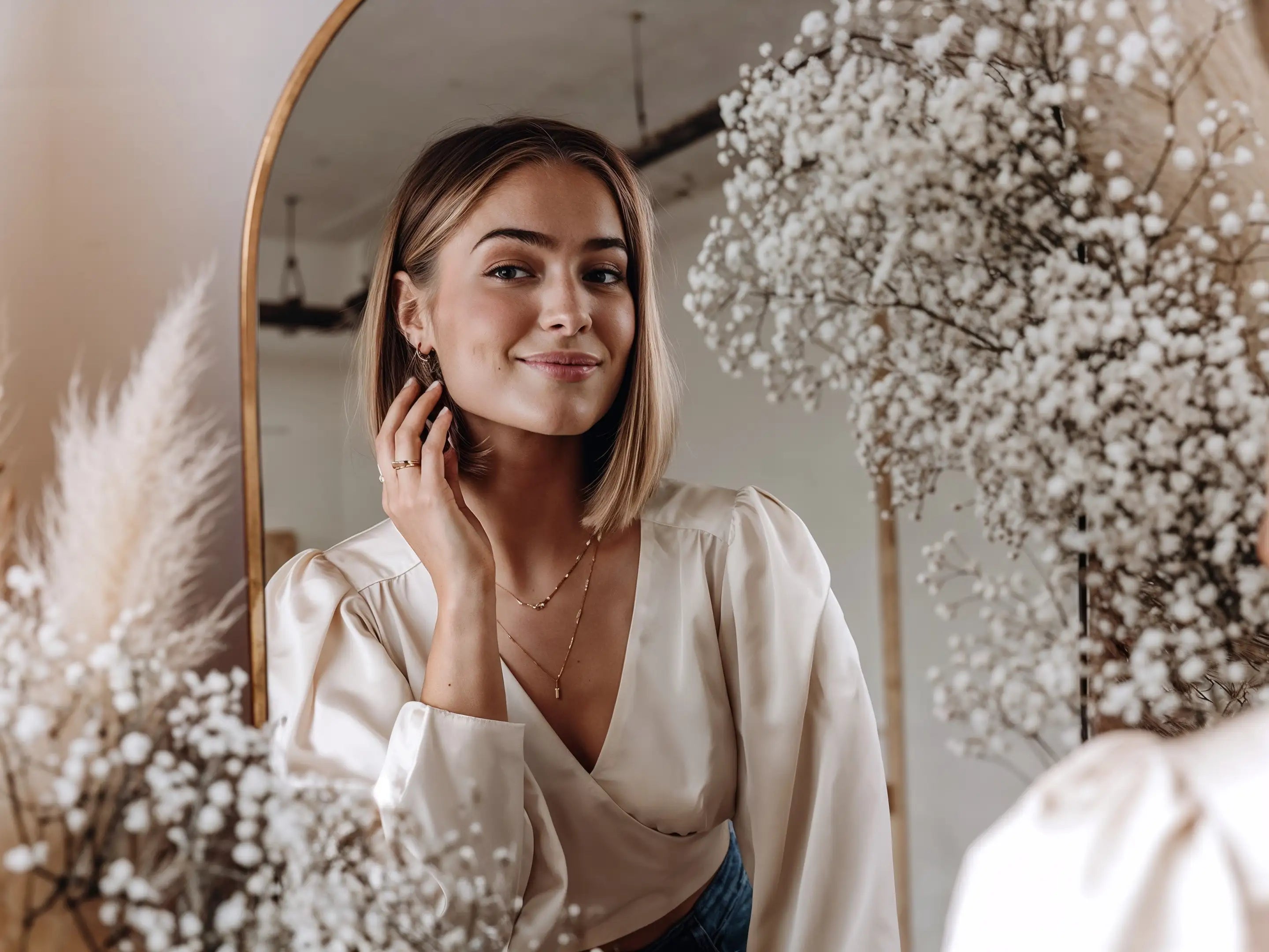 Junge Frau mit goldener Halskette, Creolen und Ring von Mercy’s Jewelry, steht vor einem Spiegel neben Blumenarrangement