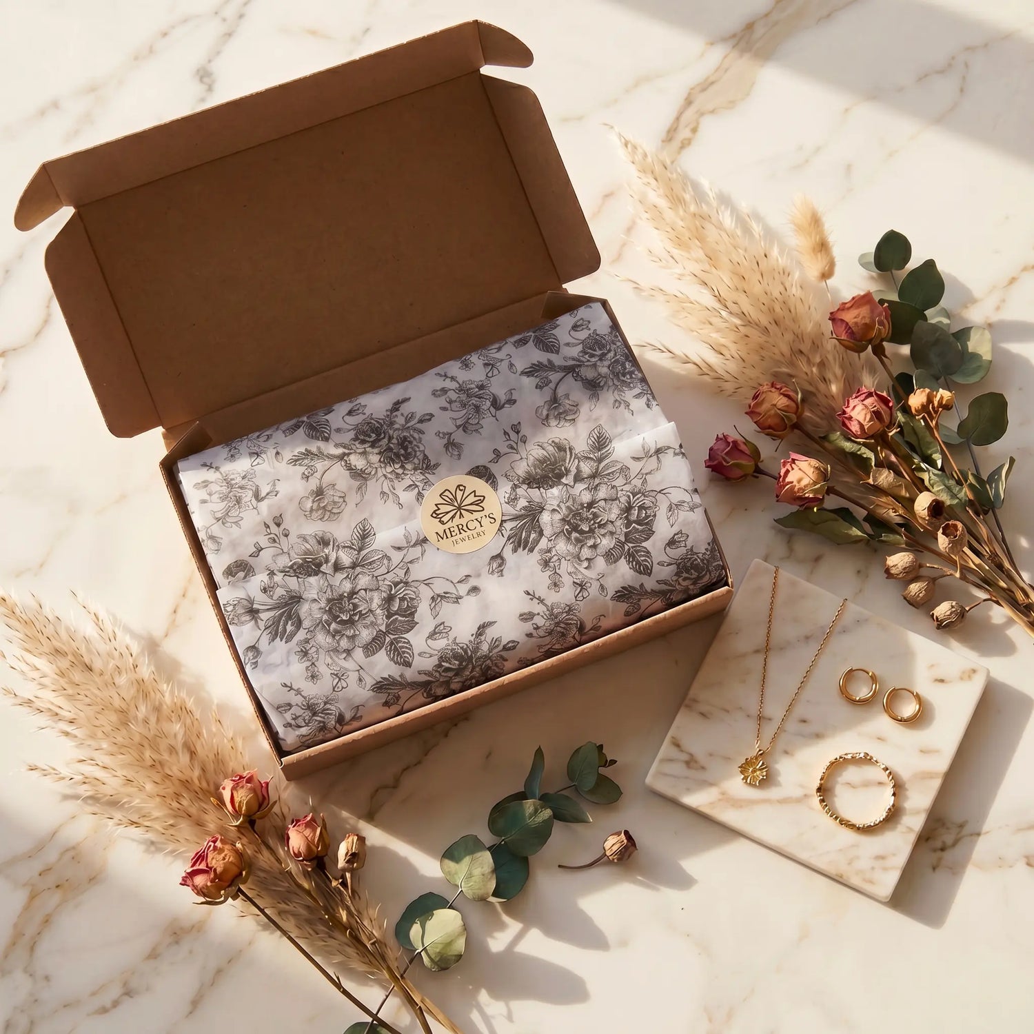 MERCY'S JEWELRY geöffnete Versandbox mit floralem Seidenpapier und goldenem Logo-Siegel, umgeben von getrockneten Rosen und Pampasgras auf Marmortisch
