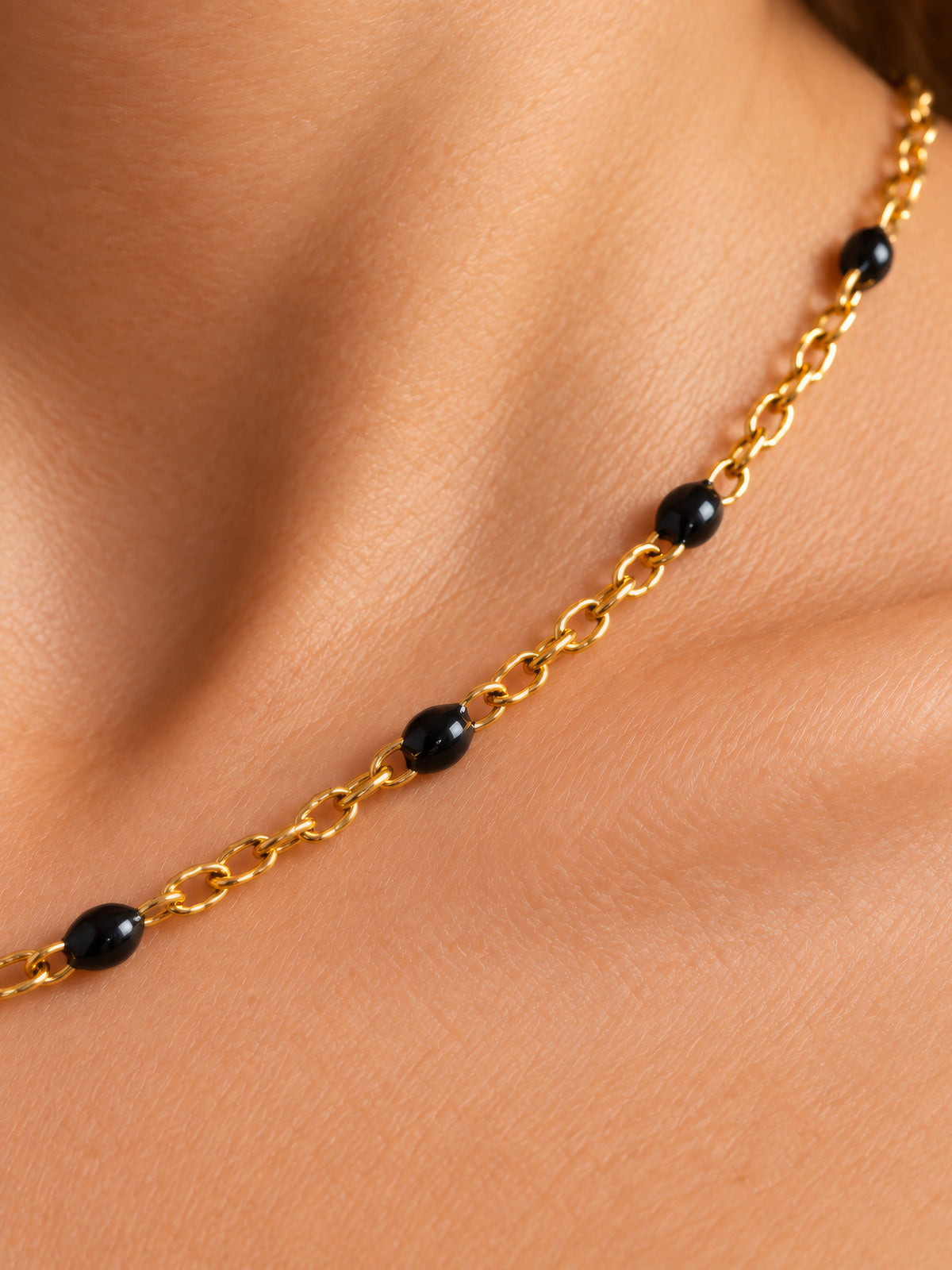 kettenglieder-detail.jpgNahaufnahme der Nivra mit glänzenden schwarzen Perlen und ineinandergreifenden goldfarbenen Kettengliedern auf der Haut.