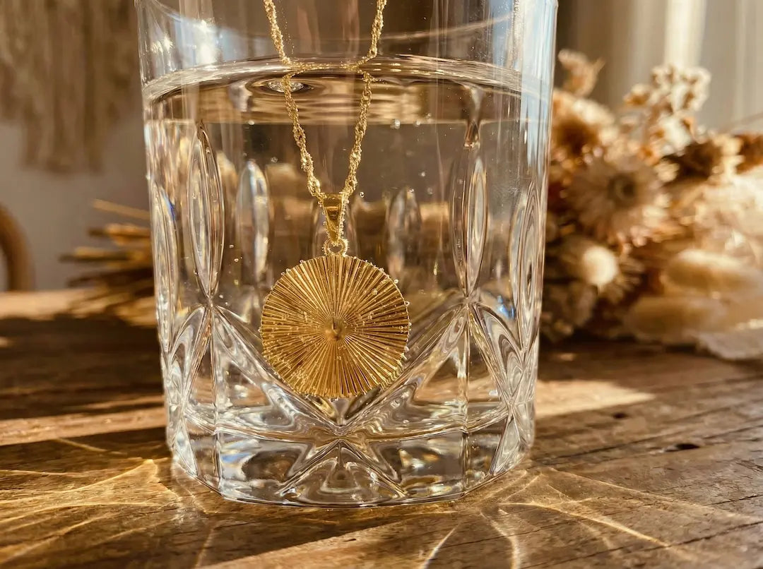 Ästhetische Nahaufnahme einer goldenen MERCYS Halskette mit rundem Sonnen-Anhänger, präsentiert in einem Glas Wasser mit Lichtreflexionen auf bymercy.de.