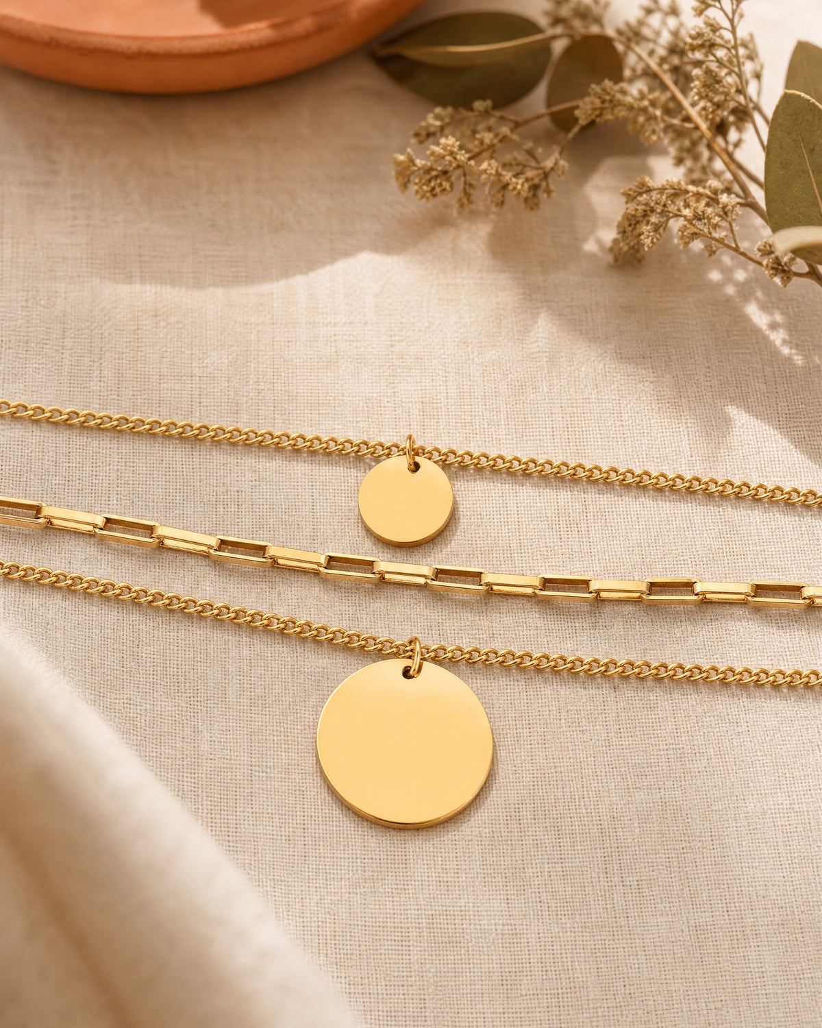 Sienna Layering Kette in Gold von MERCY'S mit drei Ketten und zwei runden Anhängern, auf hellem Leinenstoff mit Trockenblumen