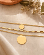 Sienna Layering Kette in Gold von MERCY'S mit drei Ketten und zwei runden Anhängern, auf hellem Leinenstoff mit Trockenblumen