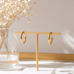 Feine Mini-Creolen aus Edelstahl 18 K vergoldet, hängen an einem goldfarbenen Schmuckständer vor hellem Hintergrund mit Trockenblumen – Nira Mini Kreolen | Mercy’s Jewelry