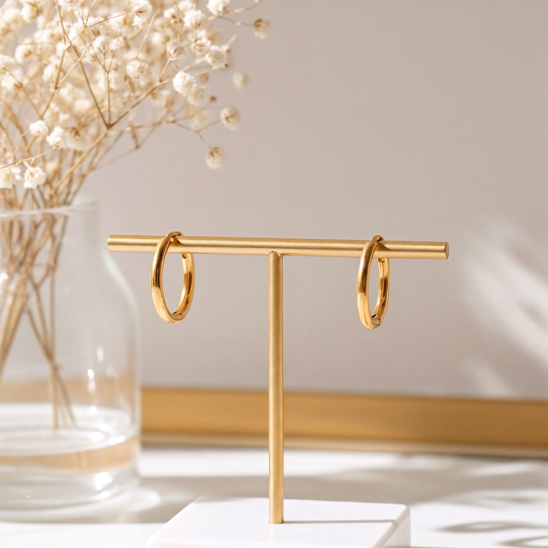 Feine Mini-Creolen aus Edelstahl 18 K vergoldet, hängen an einem goldfarbenen Schmuckständer vor hellem Hintergrund mit Trockenblumen – Nira Mini Kreolen | Mercy’s Jewelry