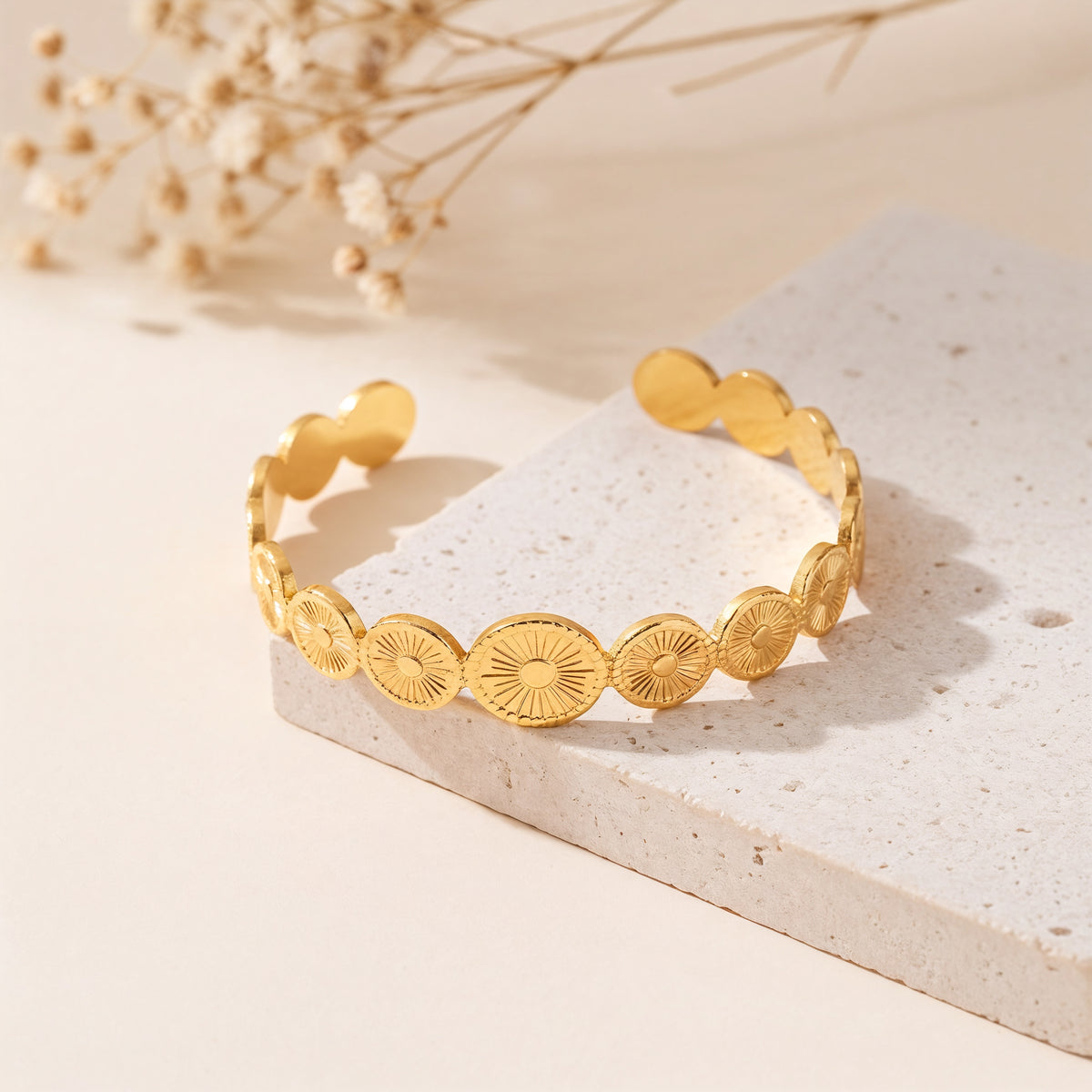Offener Armreif aus Edelstahl 18 K vergoldet mit gravierten Sonnensymbolen, liegend auf heller Steinplattform mit Trockenblumen im Hintergrund – Aurema Armreif | Mercy’s Jewelry