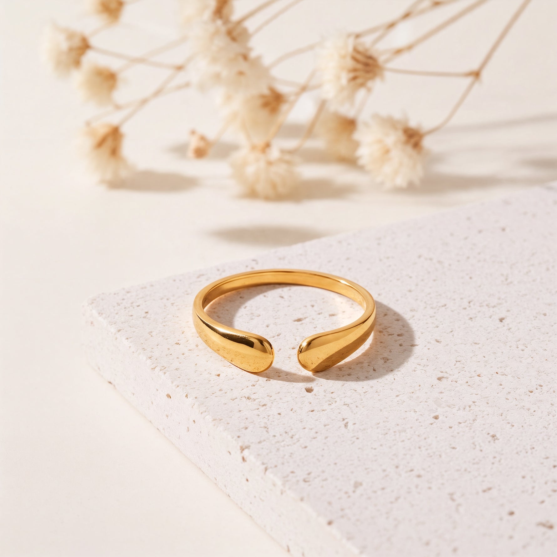 Offener Ring aus Edelstahl 18 K vergoldet, mit elegant geschwungener Tropfenform auf heller Steinplattform mit Trockenblumen im Hintergrund – Amelina Ring | Mercy’s Jewelry