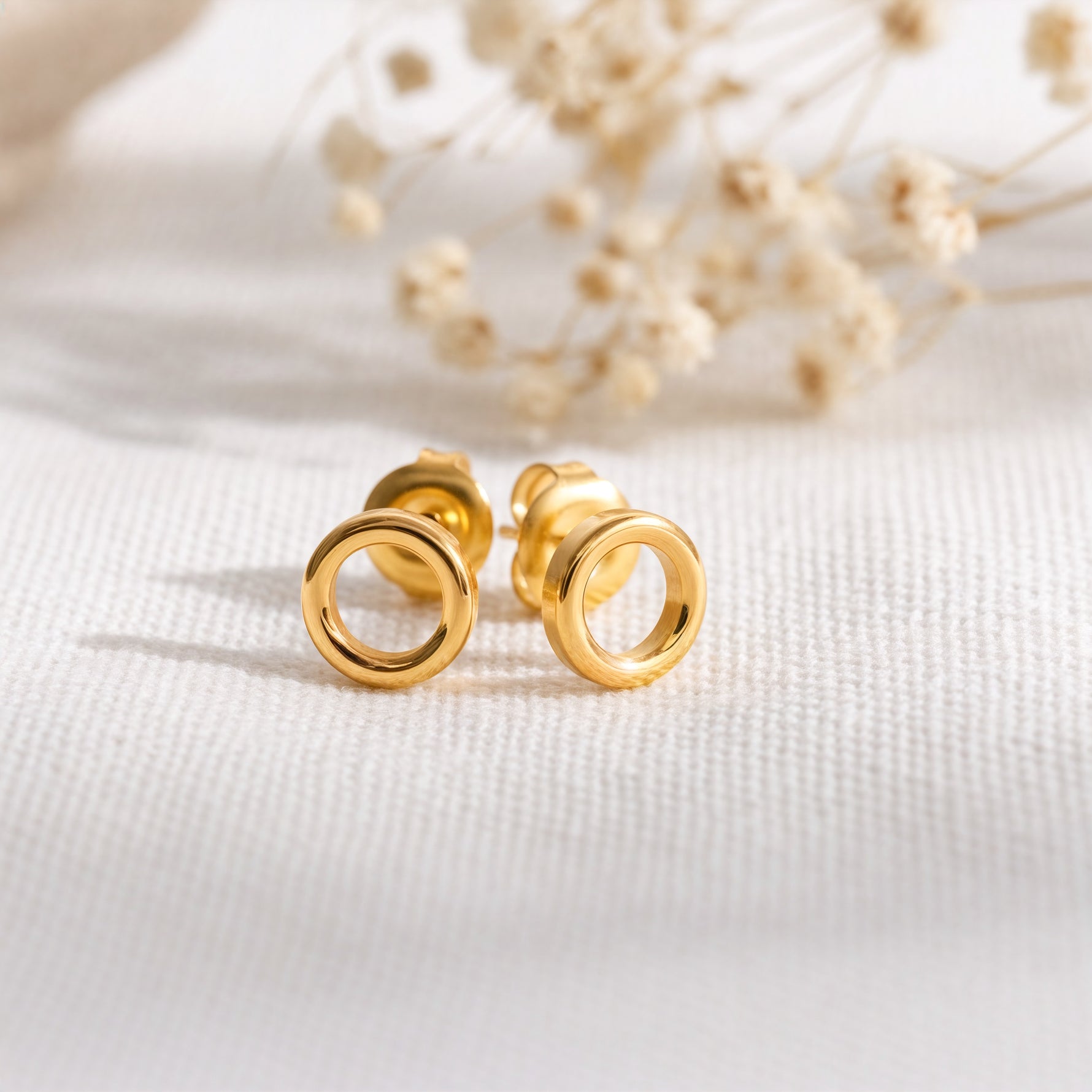 Kreisförmige Ohrstecker aus Edelstahl 18 K vergoldet, liegen auf hellem Stoff mit Trockenblumen im Hintergrund – Enoa Studs | Mercy’s Jewelry