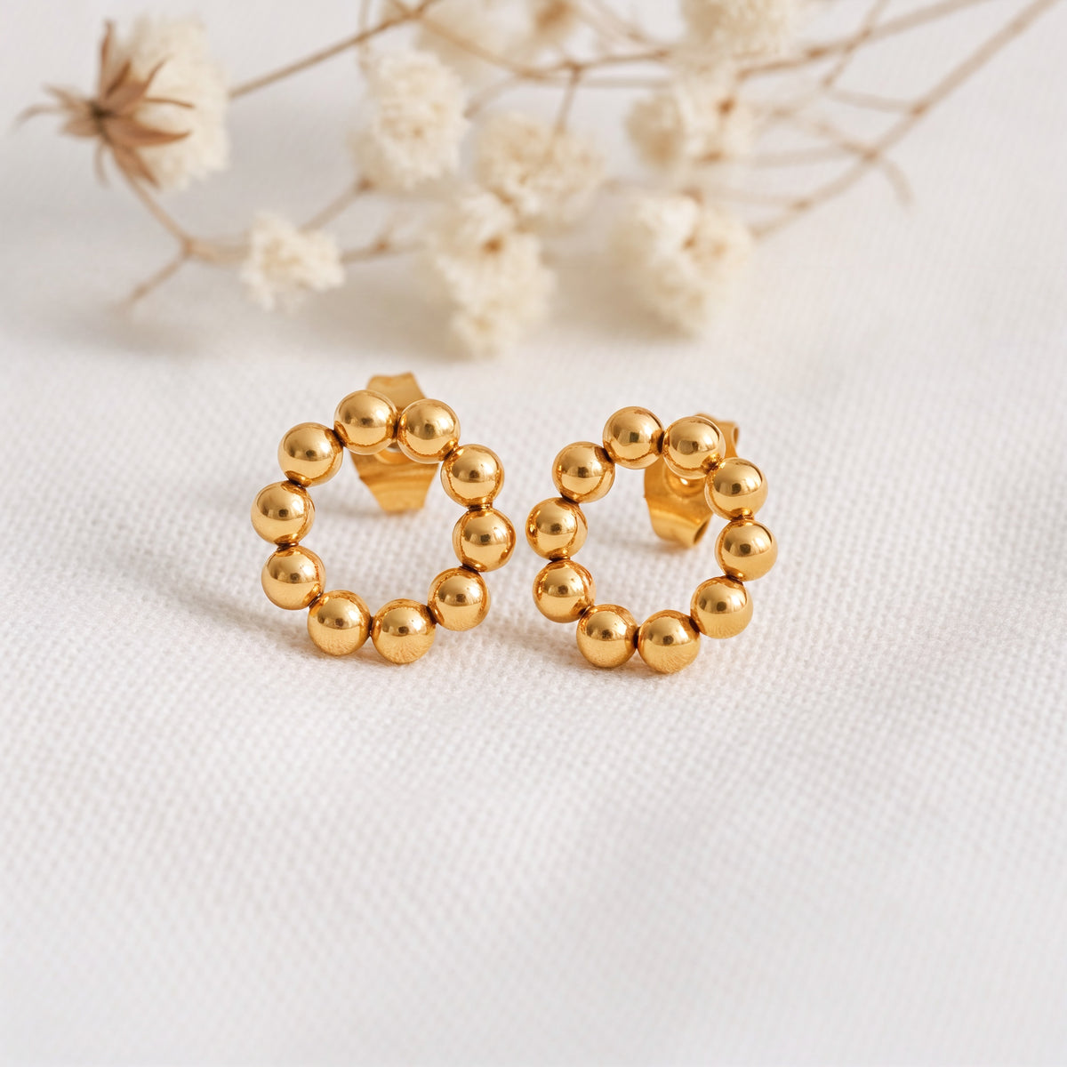 Kreisförmige Ohrstecker mit Kugel-Design aus Edelstahl 18 K vergoldet, liegen auf hellem Stoff mit Trockenblumen im Hintergrund – Orira Studs | Mercy’s Jewelry