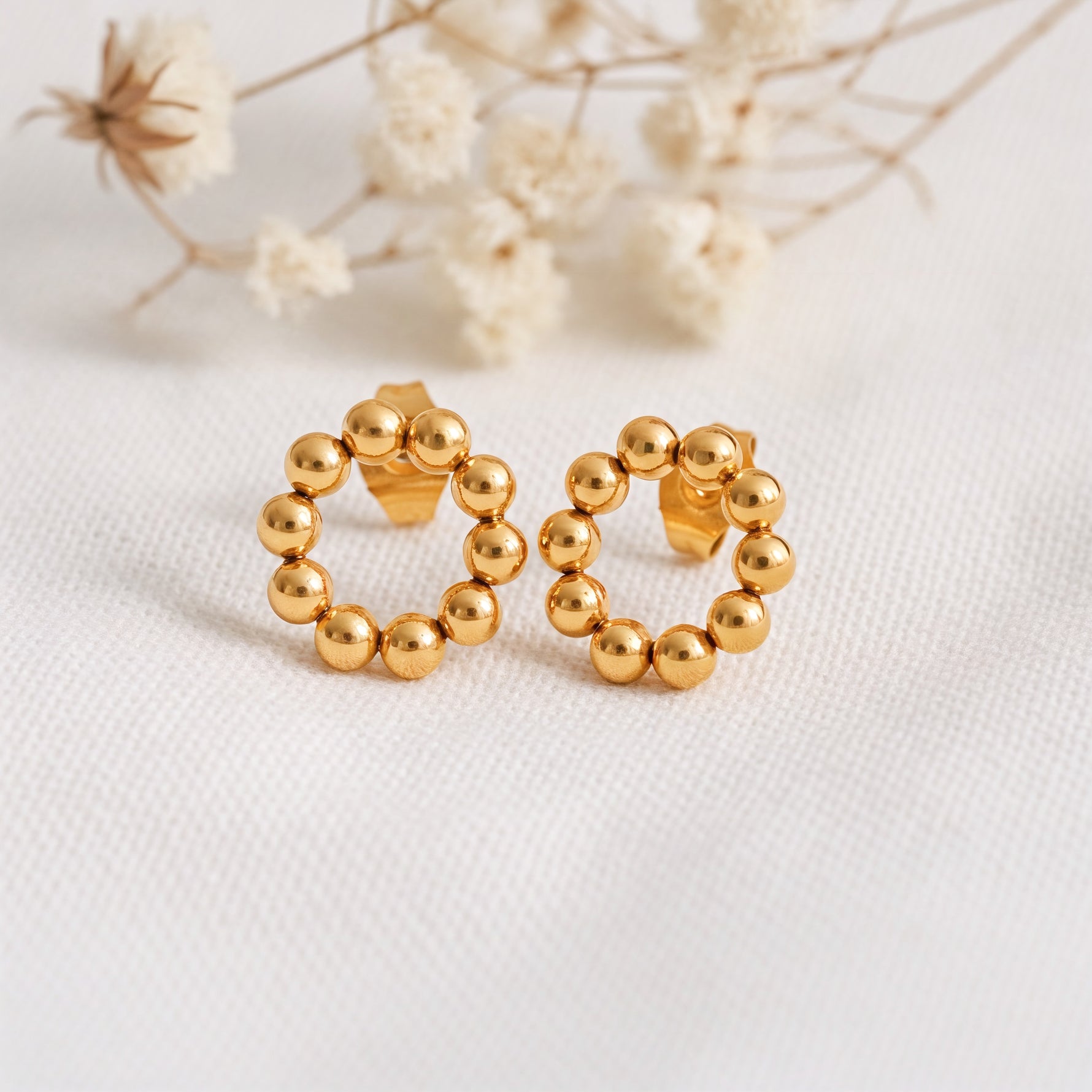 Kreisförmige Ohrstecker mit Kugel-Design aus Edelstahl 18 K vergoldet, liegen auf hellem Stoff mit Trockenblumen im Hintergrund – Orira Studs | Mercy’s Jewelry