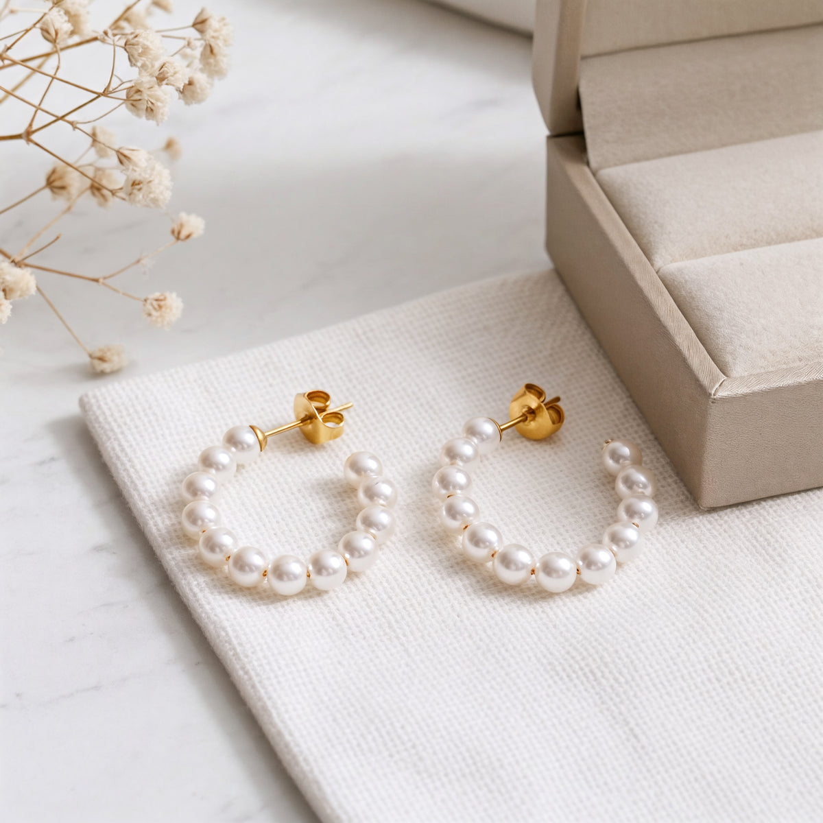 Perlen-Creolen aus Edelstahl 18 K vergoldet mit weißen runden Perlen, liegen auf hellem Stoff neben Schmuckbox und Trockenblumen – Ysira Creolen | Mercy’s Jewelry