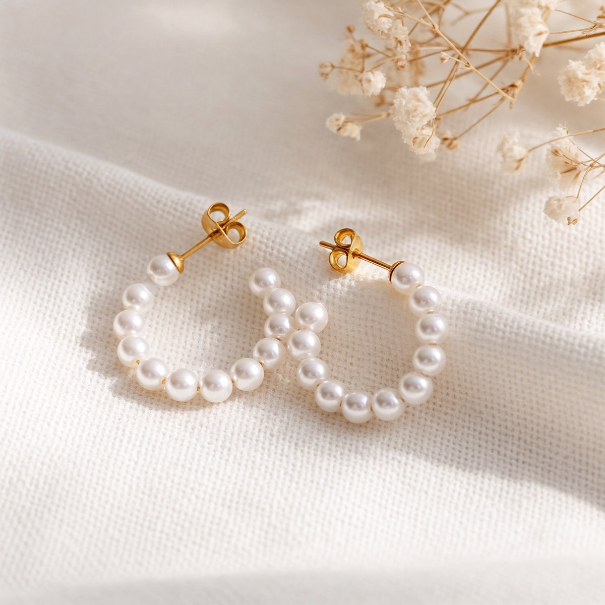 Perlen-Creolen aus Edelstahl 18 K vergoldet mit weißen runden Perlen, liegen auf hellem Stoff mit Trockenblumen im Hintergrund – Ysira Creolen | Mercy’s Jewelry