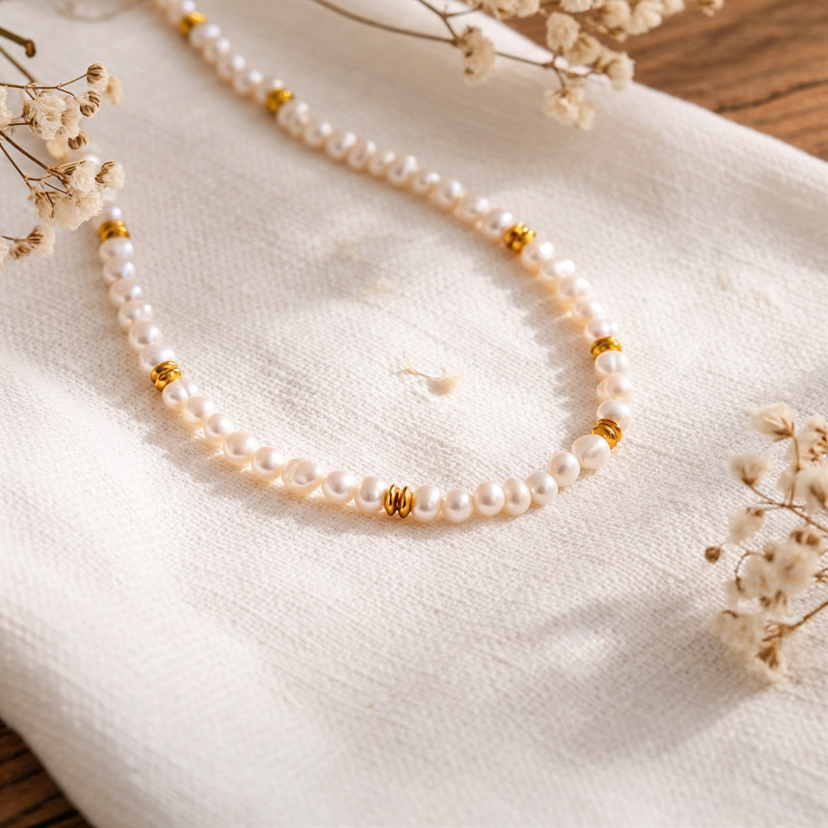 Perlenhalskette mit goldenen Zwischenelementen aus Edelstahl 18 K vergoldet, liegt auf weißem Stoff mit Trockenblumen im Hintergrund – Orielle Perlenkette | Mercy’s Jewelry