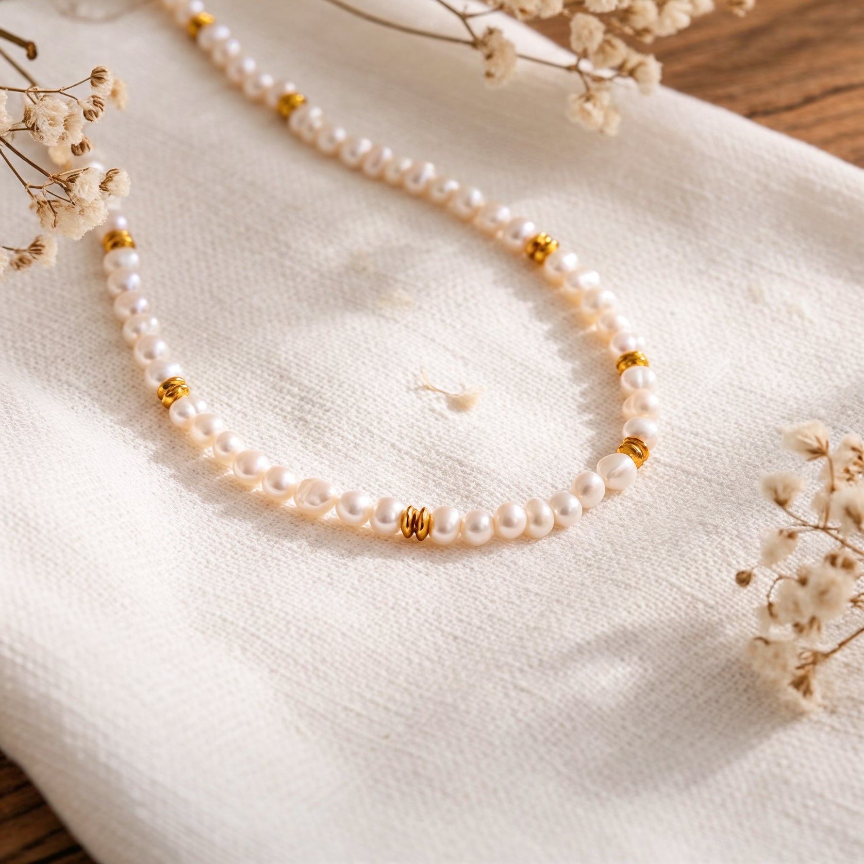 Perlenhalskette mit goldenen Zwischenelementen aus Edelstahl 18 K vergoldet, liegt auf weißem Stoff mit Trockenblumen im Hintergrund – Orielle Perlenkette | Mercy’s Jewelry