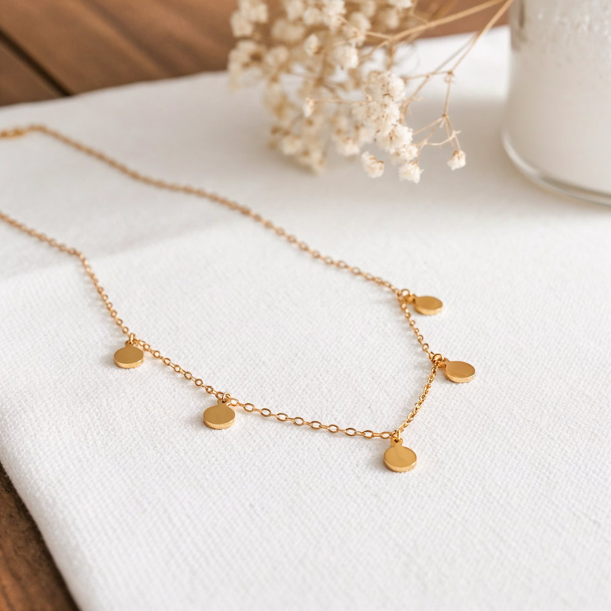 Plättchen-Halskette aus Edelstahl 18 K vergoldet, liegt auf weißem Stoff mit Trockenblumen und Vase im Hintergrund – Kaira Halskette | Mercy’s Jewelry