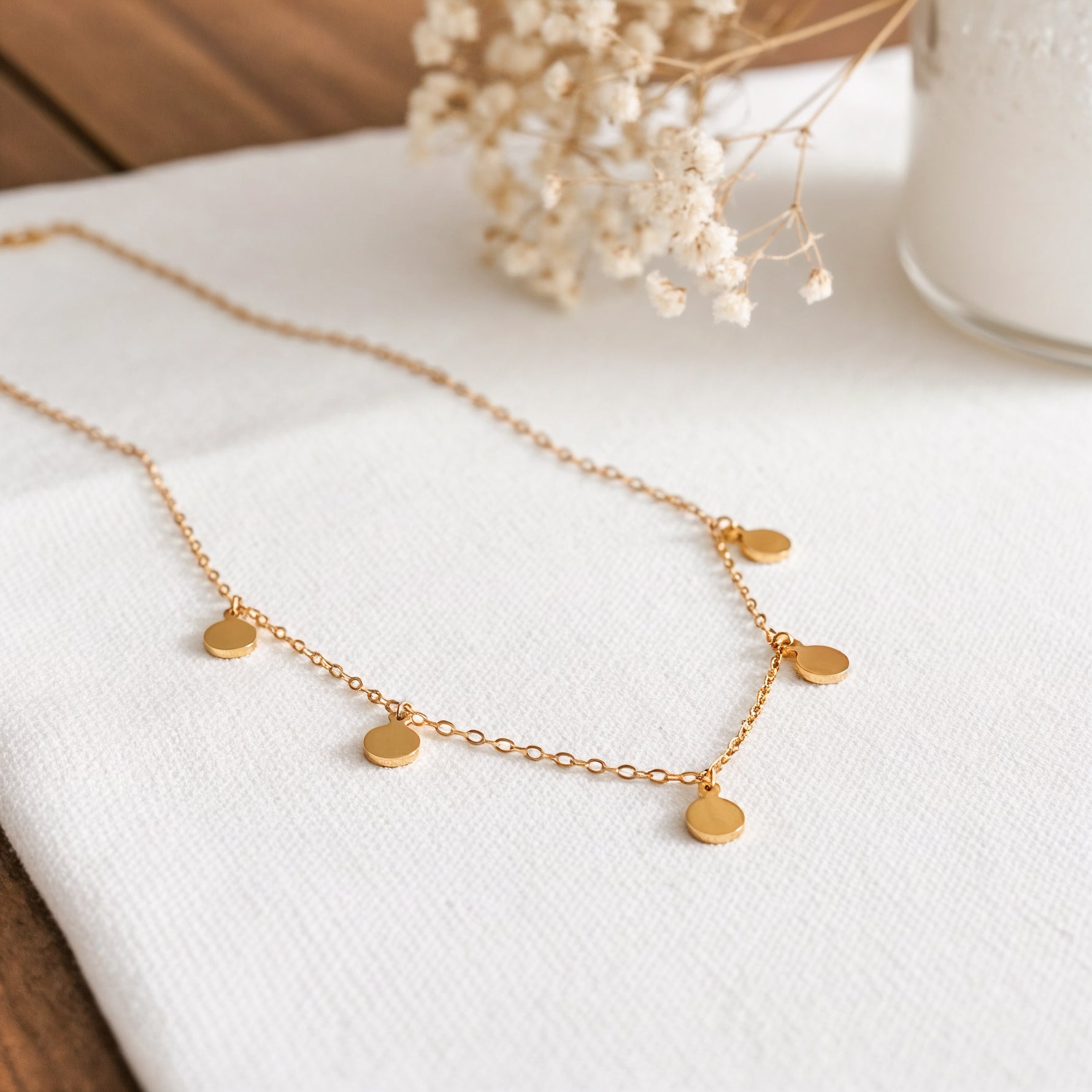Plättchen-Halskette aus Edelstahl 18 K vergoldet, liegt auf weißem Stoff mit Trockenblumen und Vase im Hintergrund – Kaira Halskette | Mercy’s Jewelry