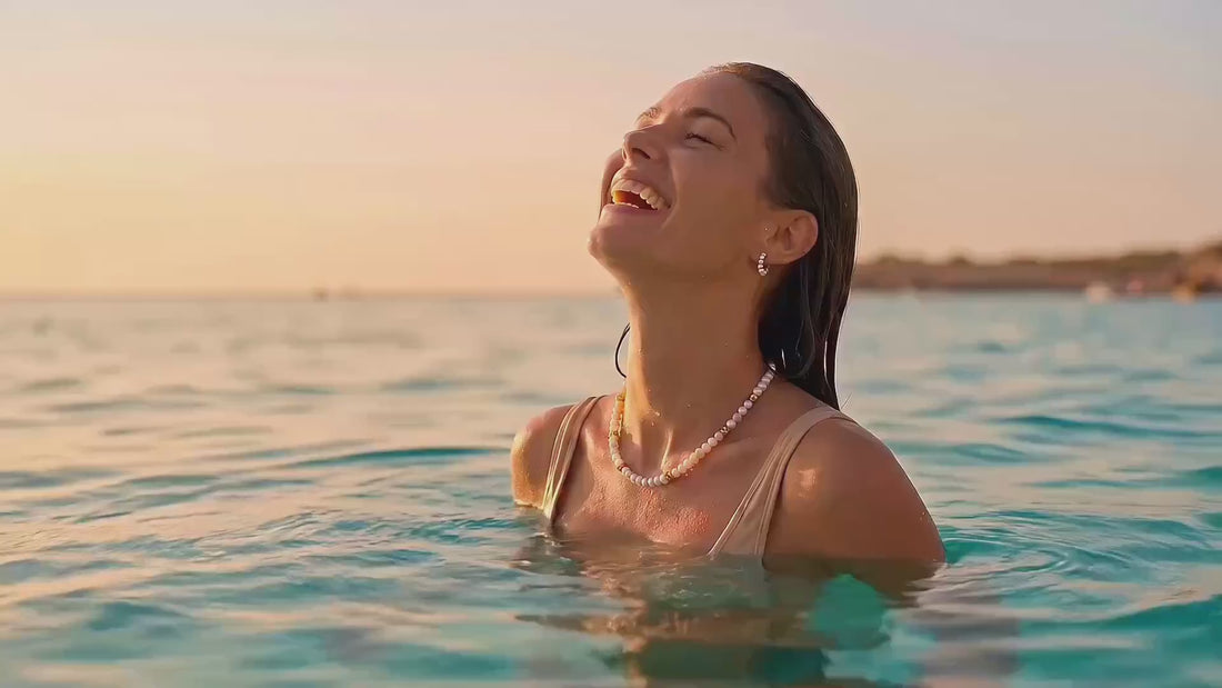 Lächelnde Frau mit wasserfester Perlenkette und Creolen von MERCY'S im Meer bei Sonnenuntergang