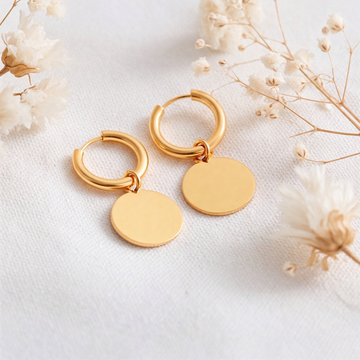 Runde Huggie-Creolen aus Edelstahl 18 K vergoldet mit flachen Plättchen-Anhängern, liegen auf hellem Stoff mit Trockenblumen im Hintergrund – Isara Huggies | Mercy’s Jewelry