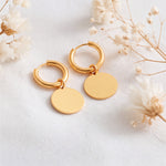 Runde Huggie-Creolen aus Edelstahl 18 K vergoldet mit flachen Plättchen-Anhängern, liegen auf hellem Stoff mit Trockenblumen im Hintergrund – Isara Huggies | Mercy’s Jewelry