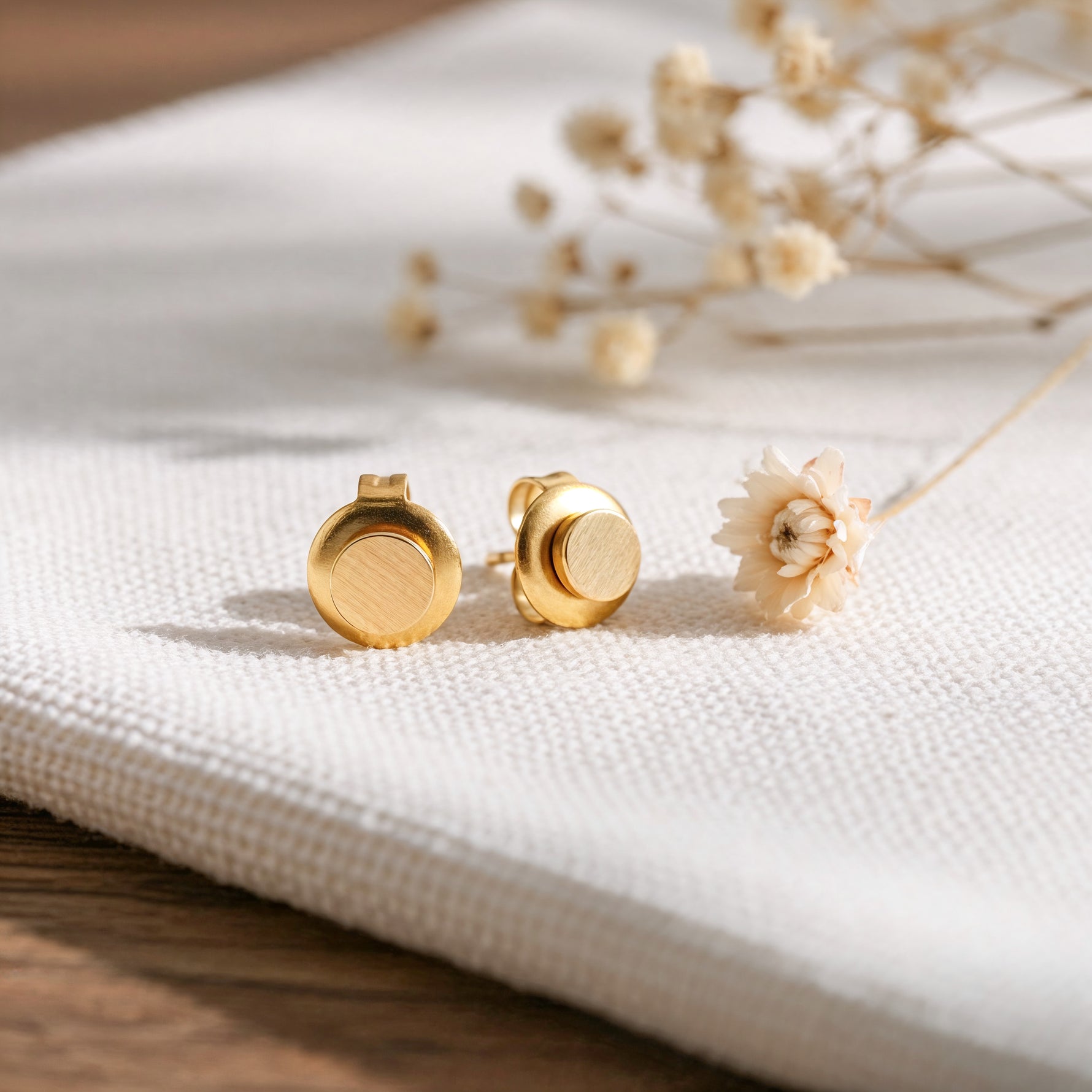 Runde Ohrstecker mit mattem und glänzendem Finish aus Edelstahl 18 K vergoldet, liegen auf hellem Stoff mit Trockenblumen im Hintergrund – Linu Studs | Mercy’s Jewelry