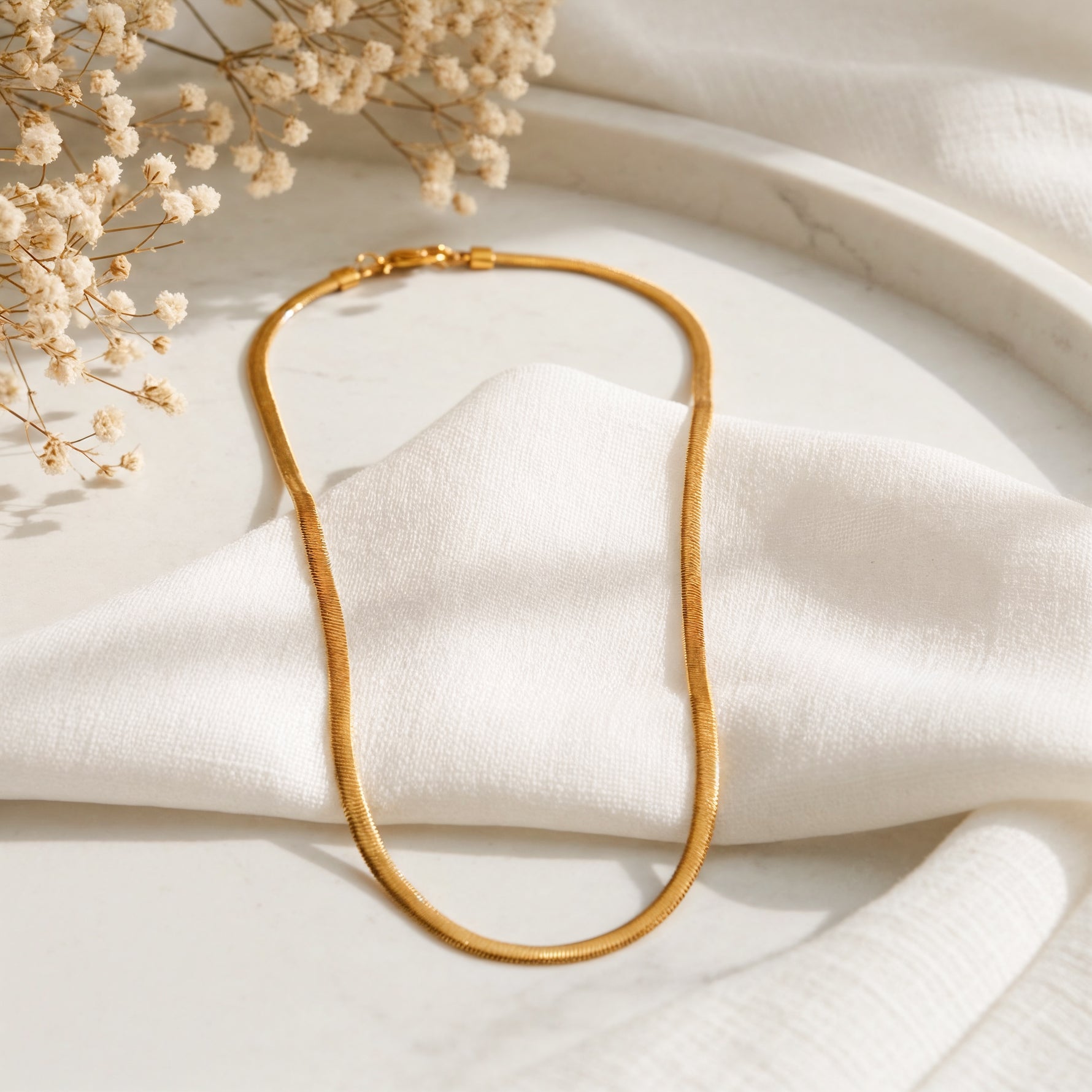 Schlangen-Halskette aus Edelstahl 18 K vergoldet, liegt auf weißem Stoff mit Trockenblumen und Marmorplatte – Soleya Halskette | Mercy’s Jewelry
