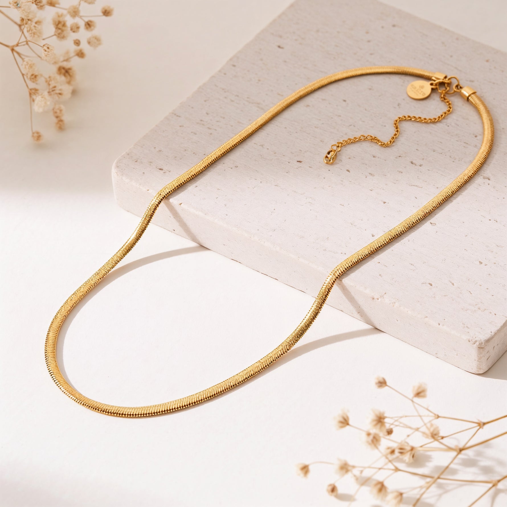 Schlangen-Halskette aus Edelstahl 18 K vergoldet, liegt auf heller Steinplatte mit Trockenblumen – Soleya Halskette | Mercy’s Jewelry