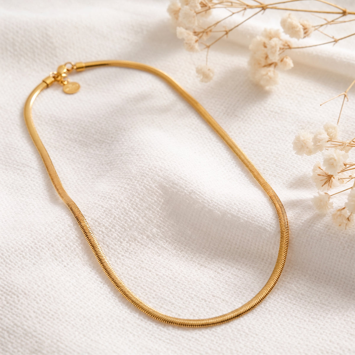Schlangen-Halskette aus Edelstahl 18 K vergoldet, liegt auf hellem Stoff mit Trockenblumen im Hintergrund – Soleya Halskette | Mercy’s Jewelry