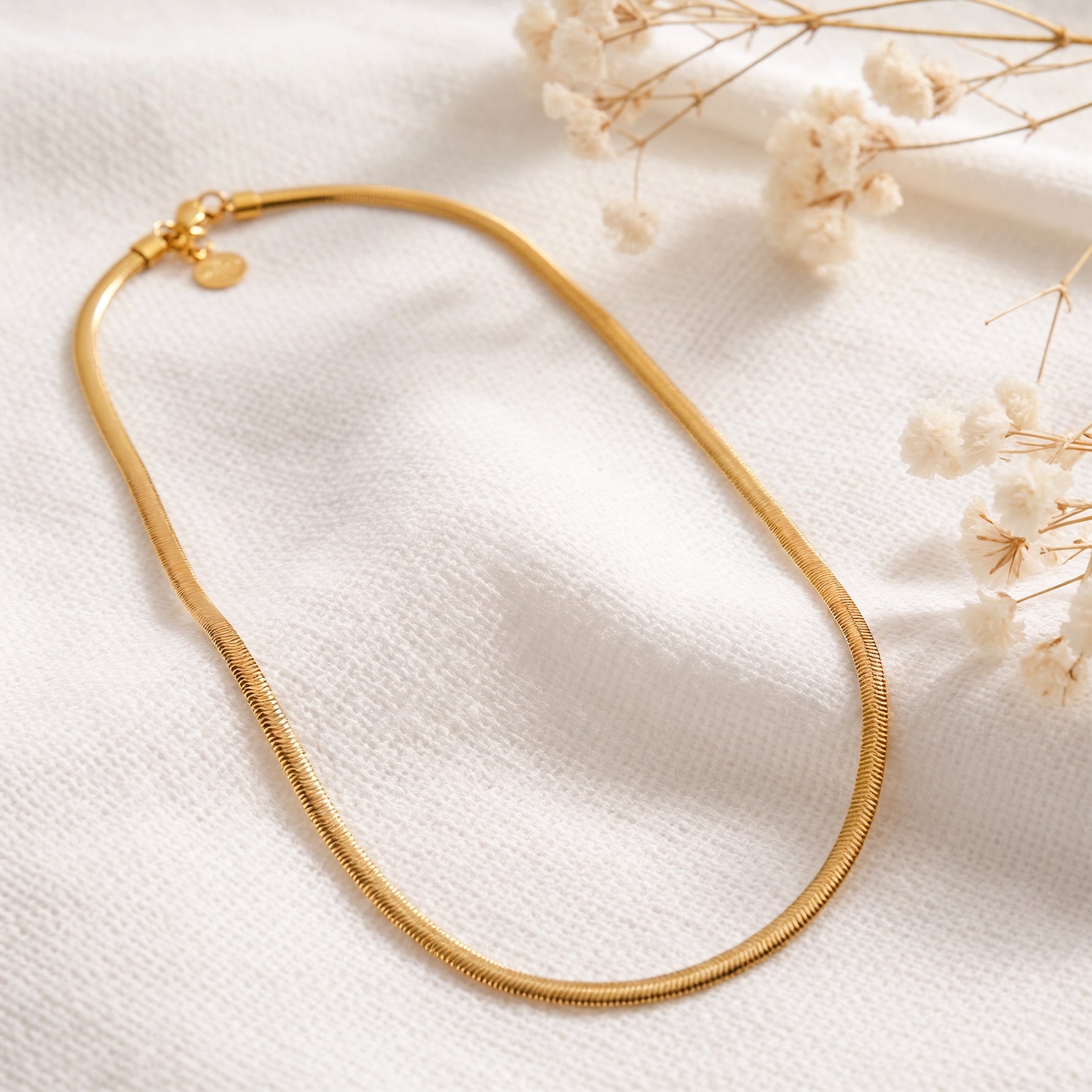 Schlangen-Halskette aus Edelstahl 18 K vergoldet, liegt auf hellem Stoff mit Trockenblumen im Hintergrund – Soleya Halskette | Mercy’s Jewelry