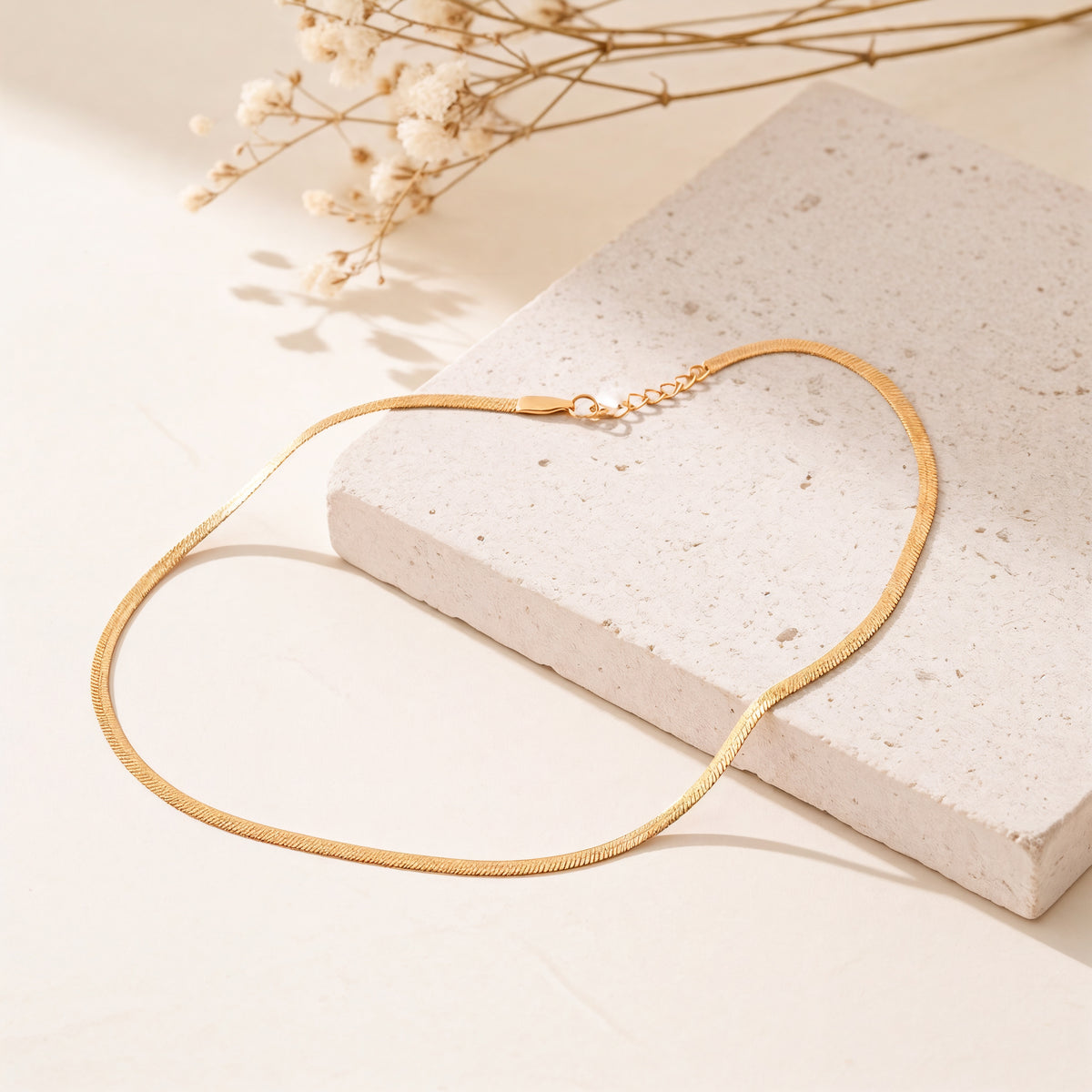 Schlangenkette aus Edelstahl 18 K vergoldet, liegt auf heller Steinplatte mit getrockneten Blumen im Hintergrund – Velira Halskette | Mercy’s Jewelry