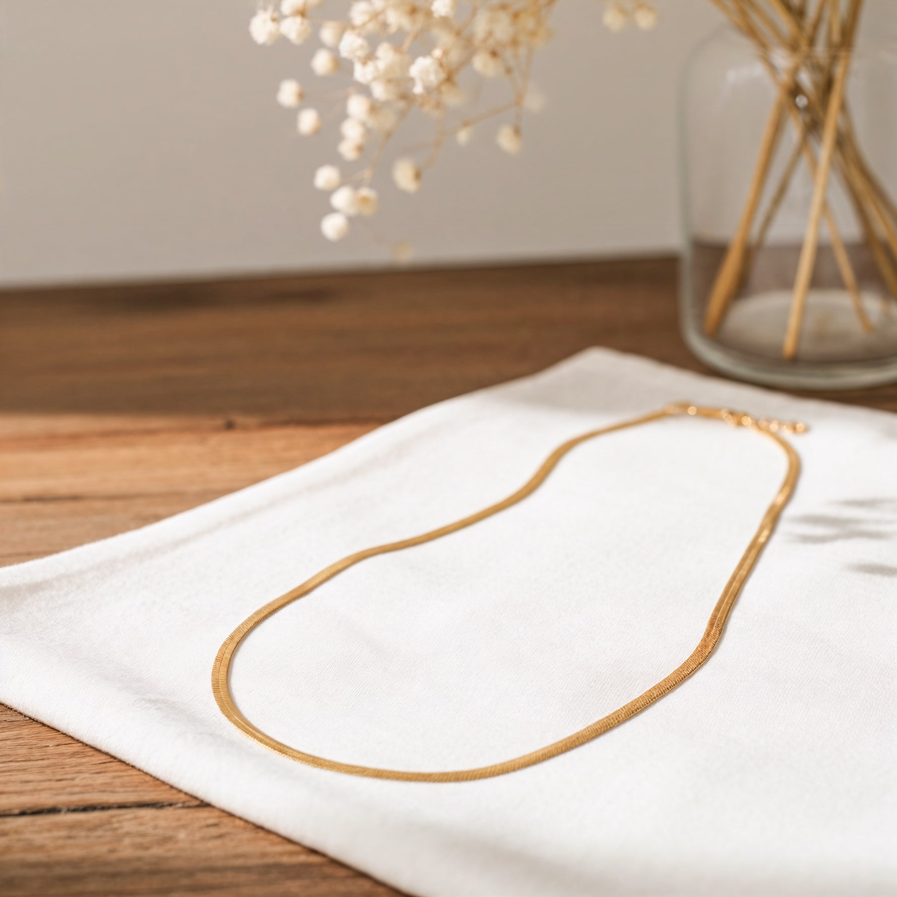 Schlangenkette aus Edelstahl 18 K vergoldet, liegt auf weißem Stoff mit Holzhintergrund und Glasvase mit Trockenblumen – Velira Halskette | Mercy’s Jewelry