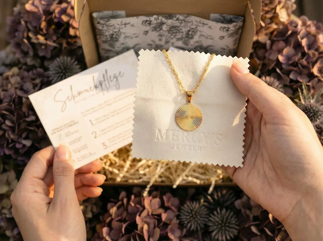 MERCY’S Jewelry Pflegeset auf bymercy.de: Weißes Poliertuch mit Logo und Schmuckpflege-Karte neben einer Goldkette.