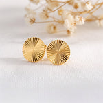 Strukturierte runde Ohrstecker aus Edelstahl 18 K vergoldet mit fein gerillter Oberfläche, liegen auf hellem Stoff mit Trockenblumen im Hintergrund – Elira Studs | Mercy’s Jewelry