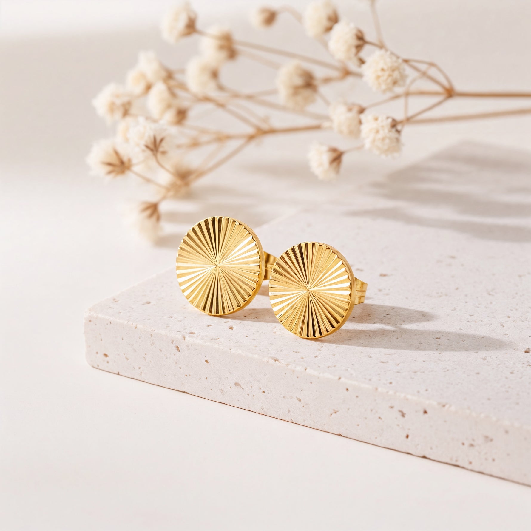 Strukturierte runde Ohrstecker aus Edelstahl 18 K vergoldet mit gerilltem Sonnendesign, liegen auf heller Steinplatte mit Trockenblumen im Hintergrund – Elira Studs | Mercy’s Jewelry