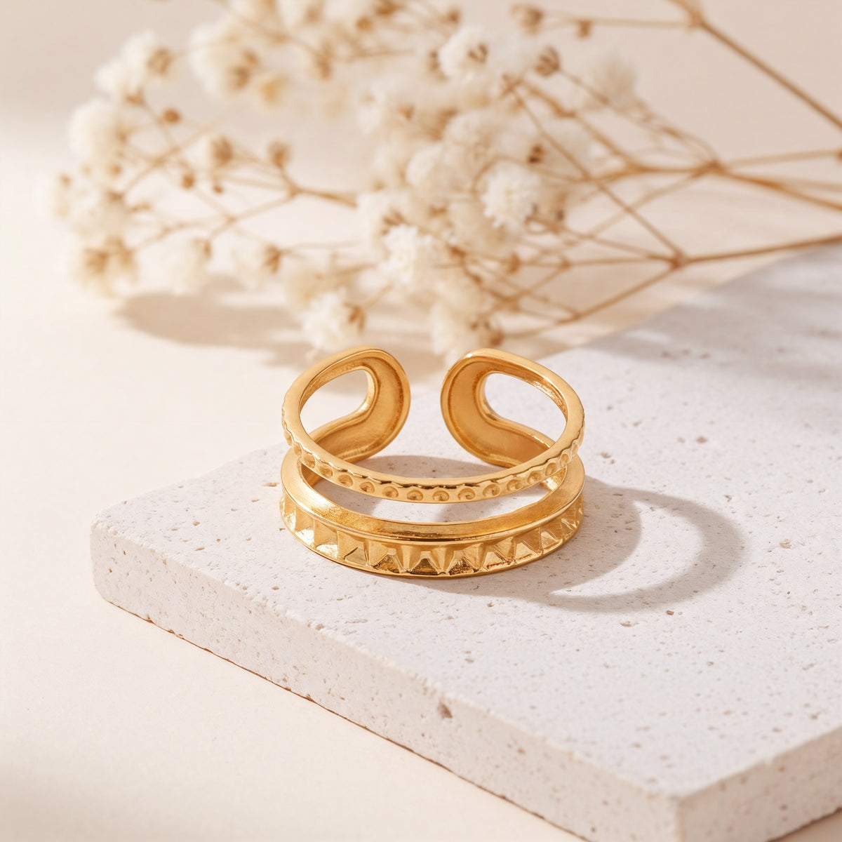 Strukturierter Doppelring aus Edelstahl 18 K vergoldet, offen und verstellbar, mit pyramidenförmigem und kugelförmigem Relief-Design auf heller Steinplattform mit Trockenblumen im Hintergrund – Sireva Ring | Mercy’s Jewelry
