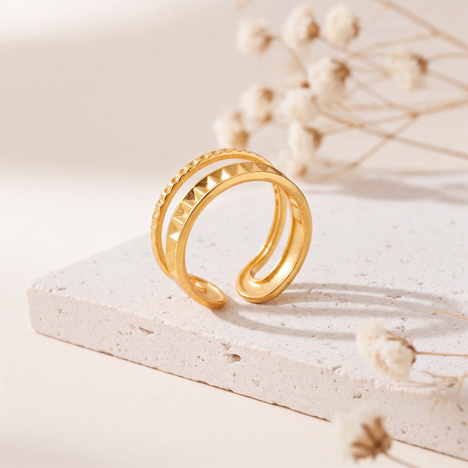 Strukturierter Doppelring aus Edelstahl 18 K vergoldet, offen und verstellbar, mit pyramidenförmigem Muster und polierter Oberfläche auf heller Steinplattform mit Trockenblumen im Hintergrund – Sireva Ring | Mercy’s Jewelry