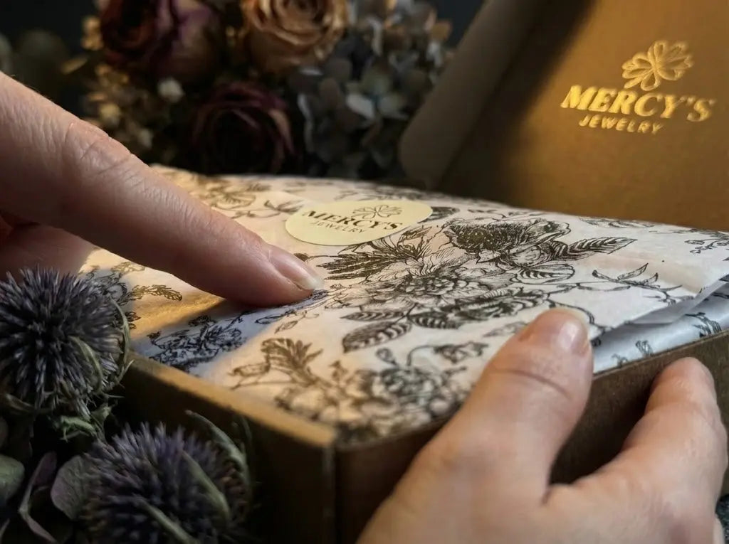 Das MERCY’S Jewelry Unboxing-Erlebnis auf bymercy.de: Hände öffnen floral bedrucktes Seidenpapier mit Logo-Sticker.