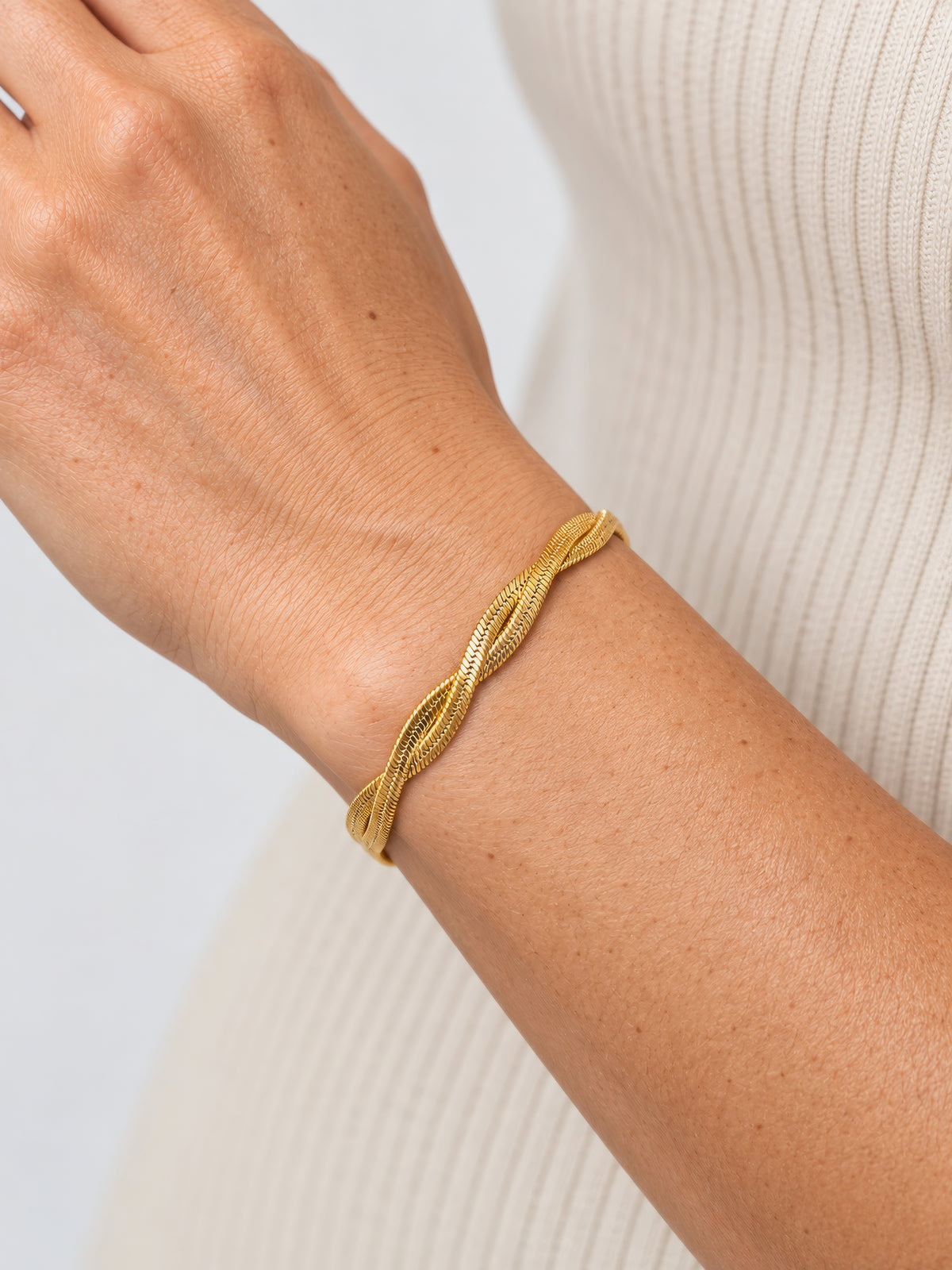 Velira Double Armband in Gold am Handgelenk, zwei gedrehte Fischgrätenstränge, Nahaufnahme vor cremefarbenem Strick