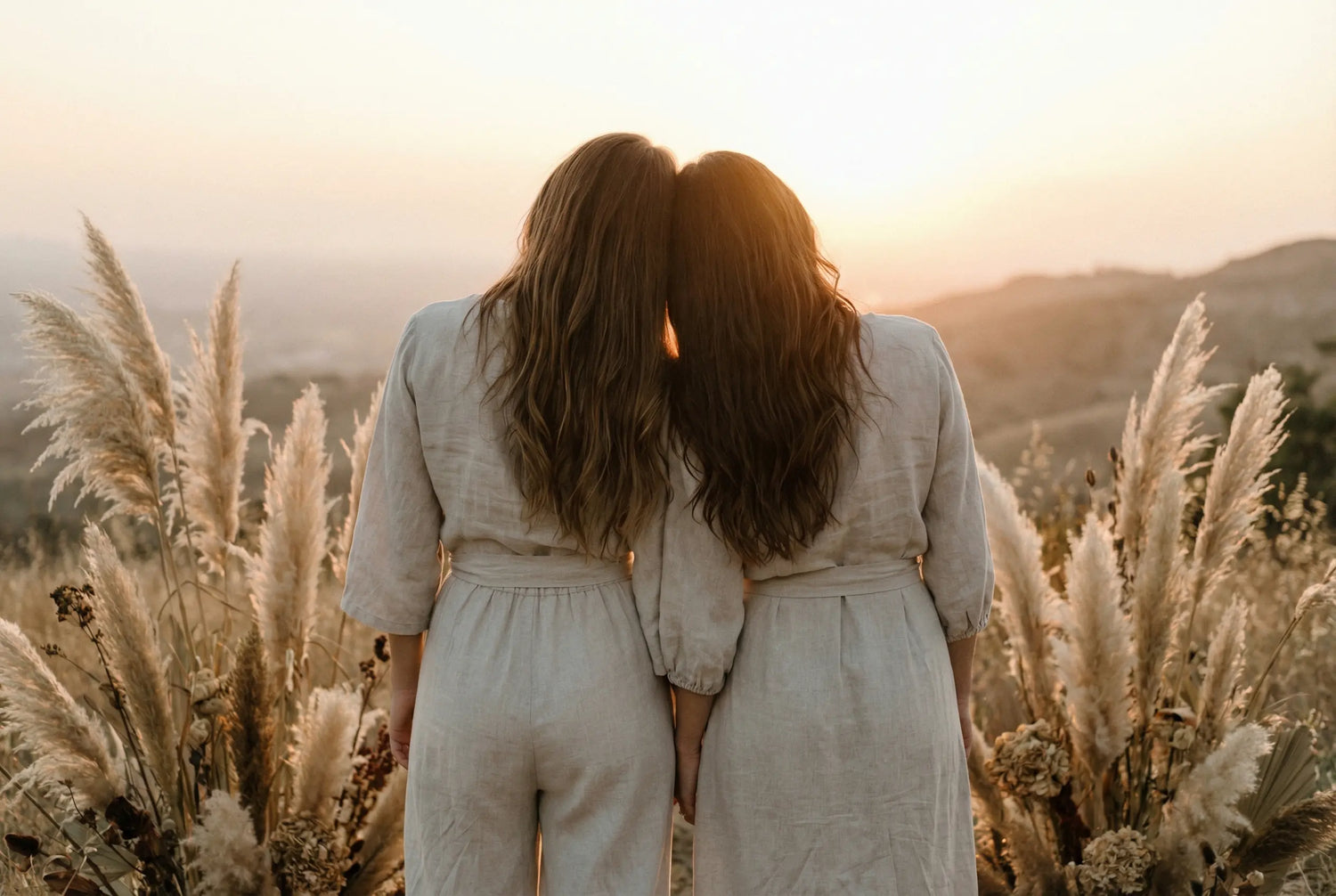 Zwei Frauen in hellen Leinenkleidern stehen nah beieinander mit dem Rücken zur Kamera und blicken auf einen warmen Sonnenuntergang, umgeben von Pampasgras.