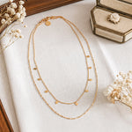 Zweireihige Halskette aus Edelstahl 18 K vergoldet mit kleinen quadratischen Anhängern, liegt auf beigem Stoff mit Trockenblumen und Schmuckbox – Zelina Layers Halskette | Mercy’s Jewelry