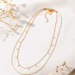 Zweireihige Halskette aus Edelstahl 18 K vergoldet mit kleinen quadratischen Anhängern, liegt auf heller Marmoroberfläche mit weißen Trockenblumen – Zelina Layers Halskette | Mercy’s Jewelry