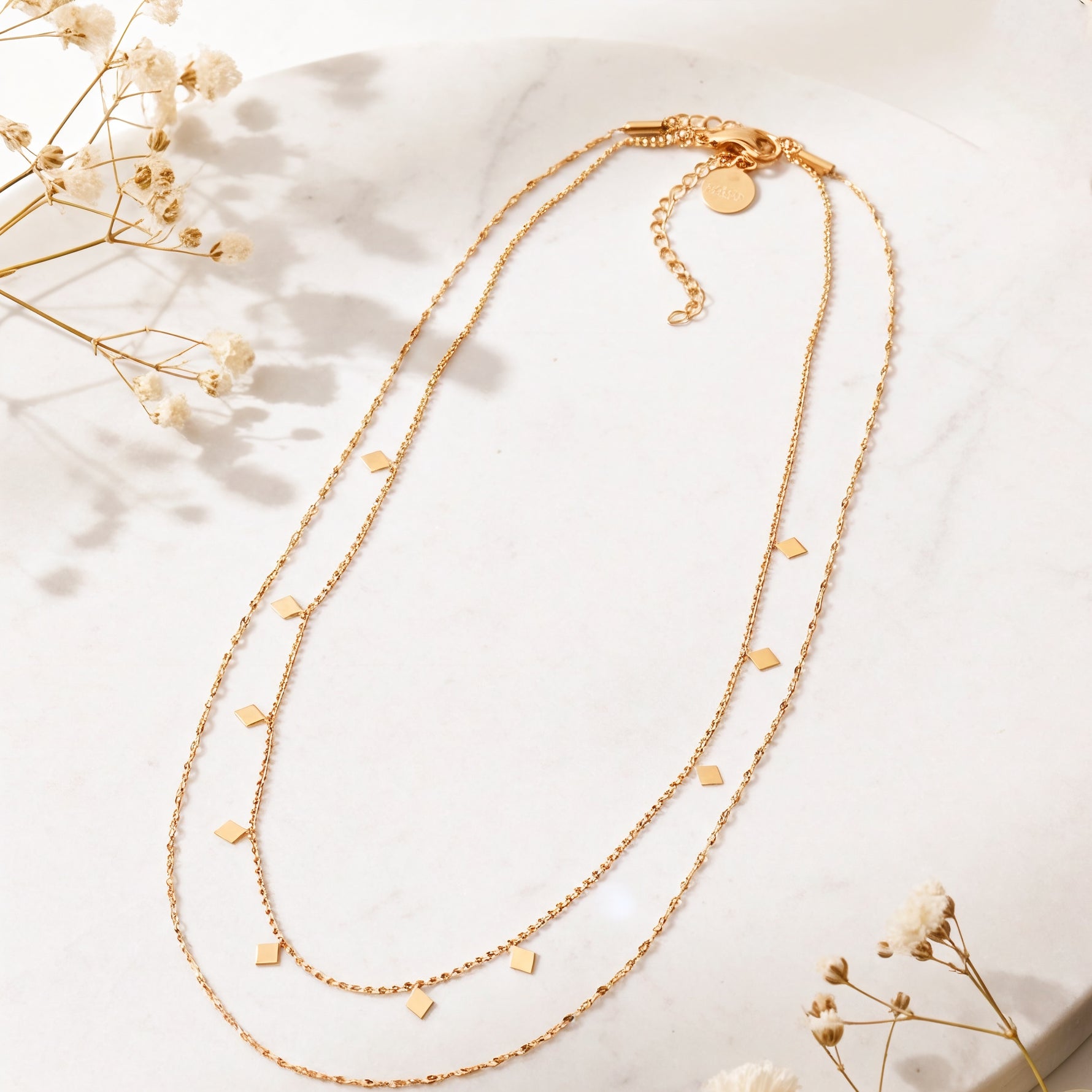 Zweireihige Halskette aus Edelstahl 18 K vergoldet mit kleinen quadratischen Anhängern, liegt auf heller Marmoroberfläche mit weißen Trockenblumen – Zelina Layers Halskette | Mercy’s Jewelry